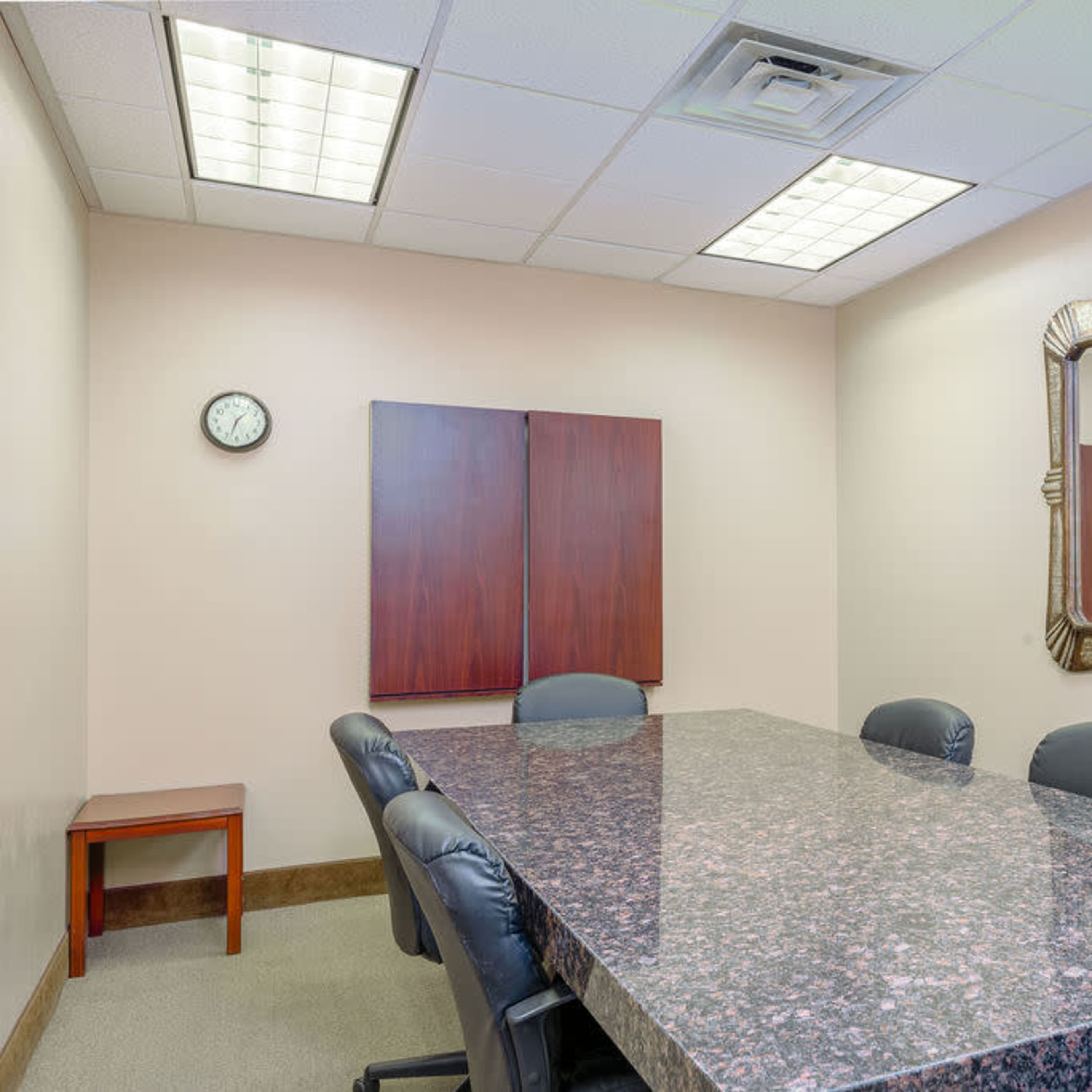 A conference room features a dark marble table surrounded by black chairs, with a clock on the wall and a large decorative mirror.