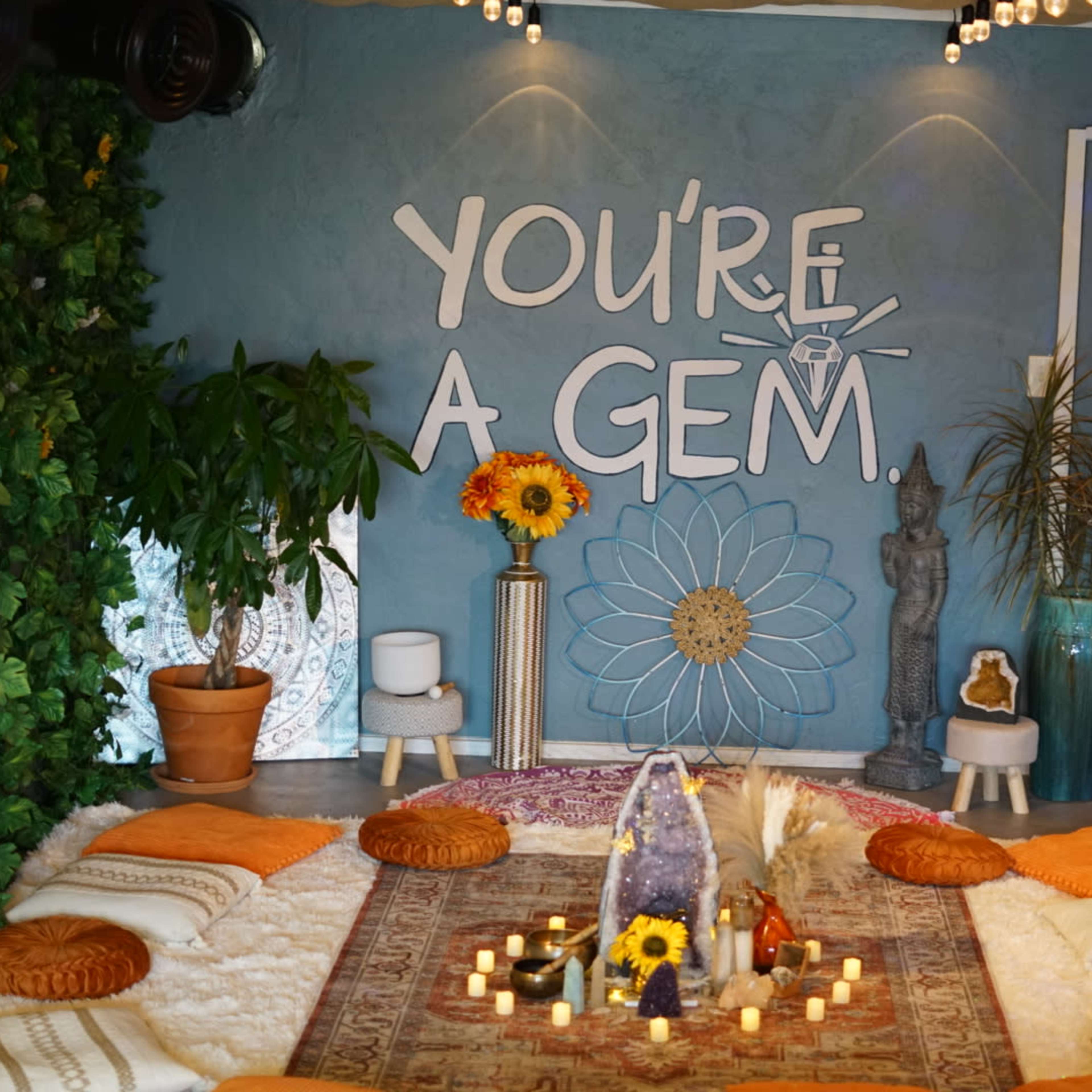 A cozy interior space features a central rug surrounded by cushions, with a decorative wall that reads "YOU'RE A GEM" and various plants and candles throughout.