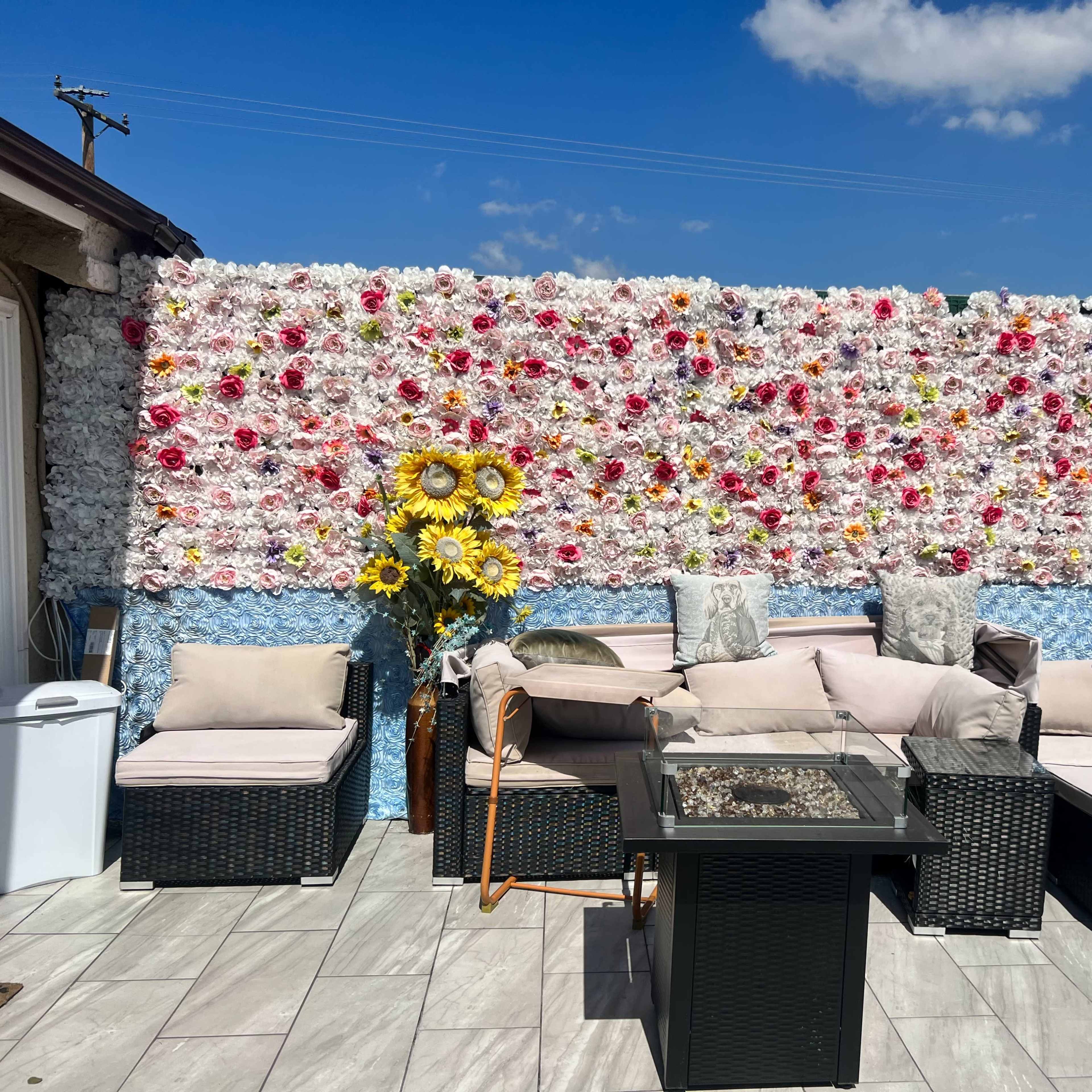 A rooftop patio with a flower-covered wall, comfortable seating arranged around a glass table, and a sunny sky in the background.