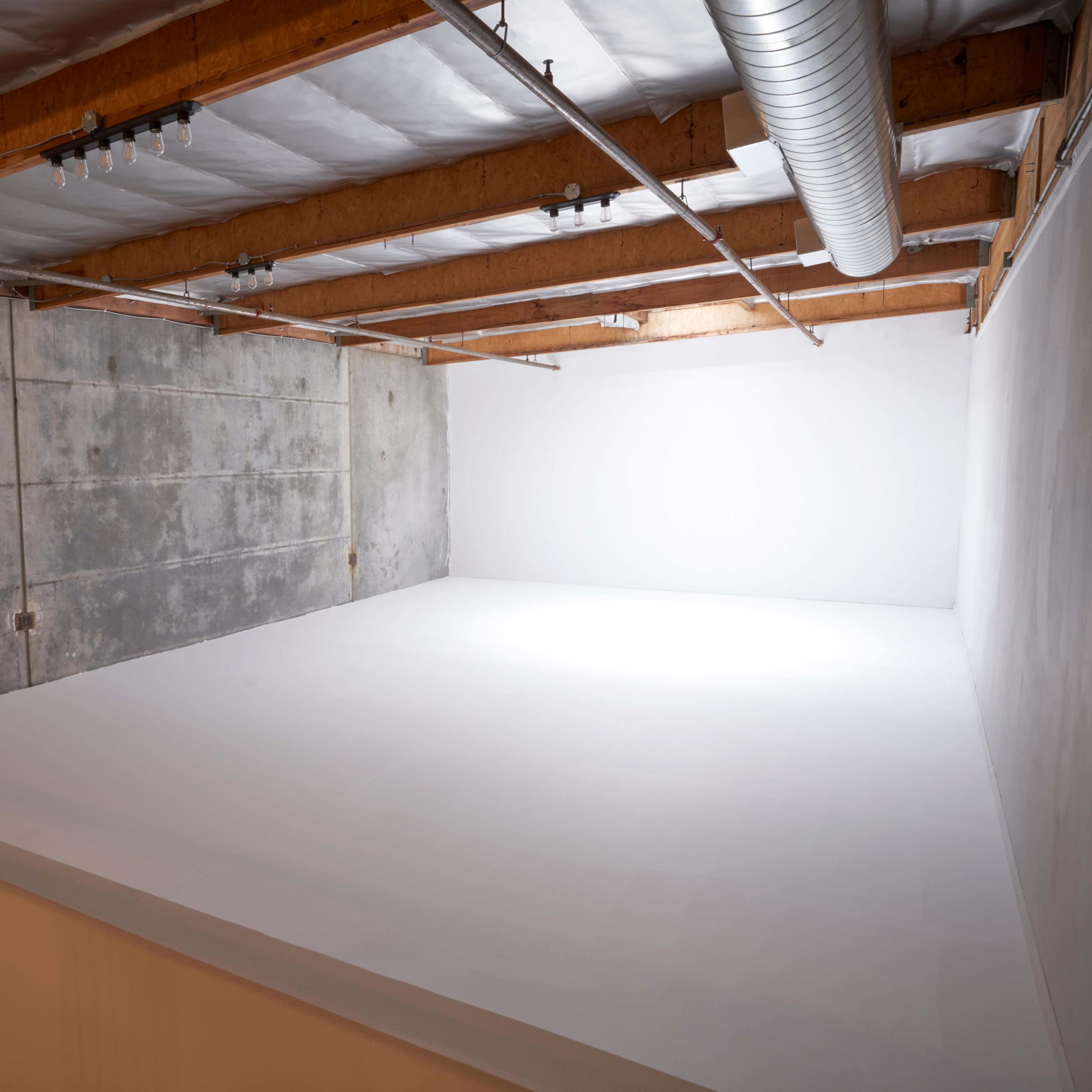 A spacious, empty studio with wooden beams and concrete walls, featuring a bright, white corner illuminated by soft light.