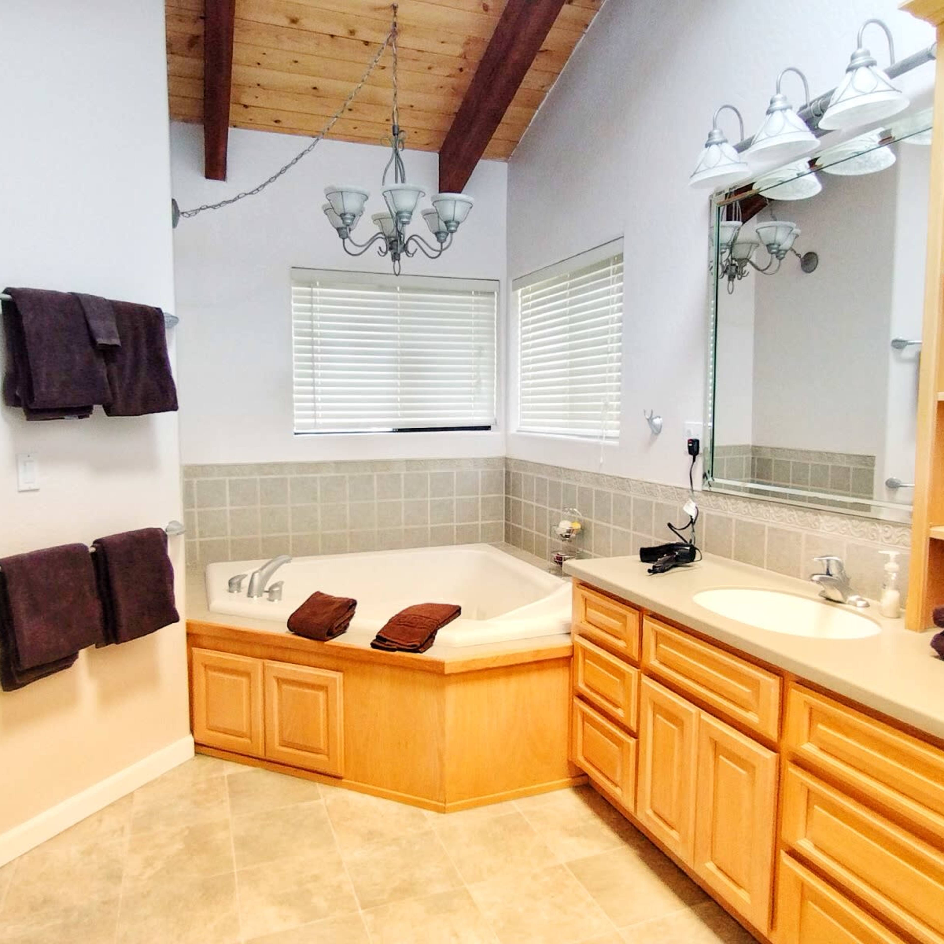 A bright bathroom features a wood-beamed ceiling, a corner jetted bathtub, a double sink vanity, and neatly arranged towels on the racks.