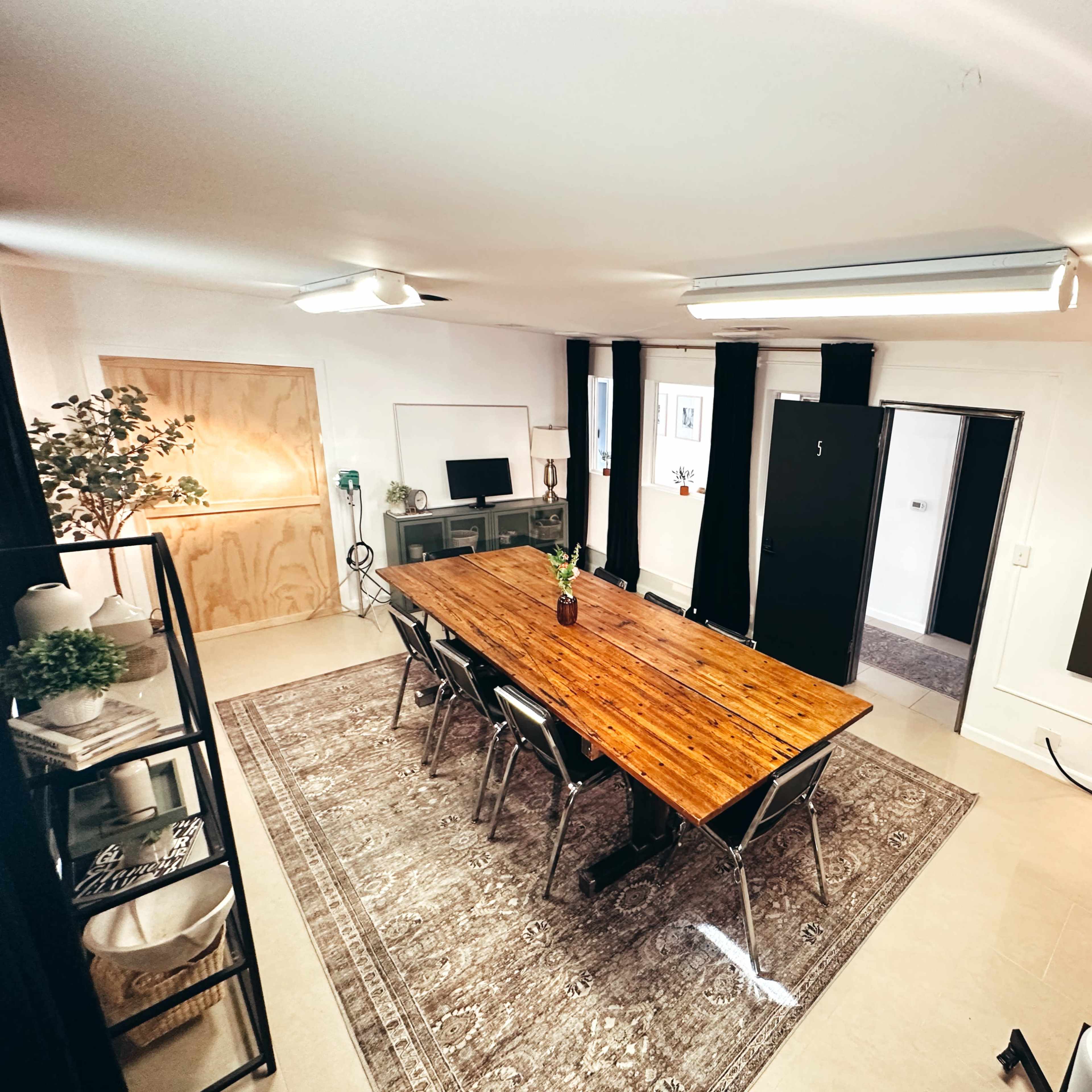 A spacious meeting room features a long wooden table surrounded by dark chairs, with a television on one wall and framed artwork on another.