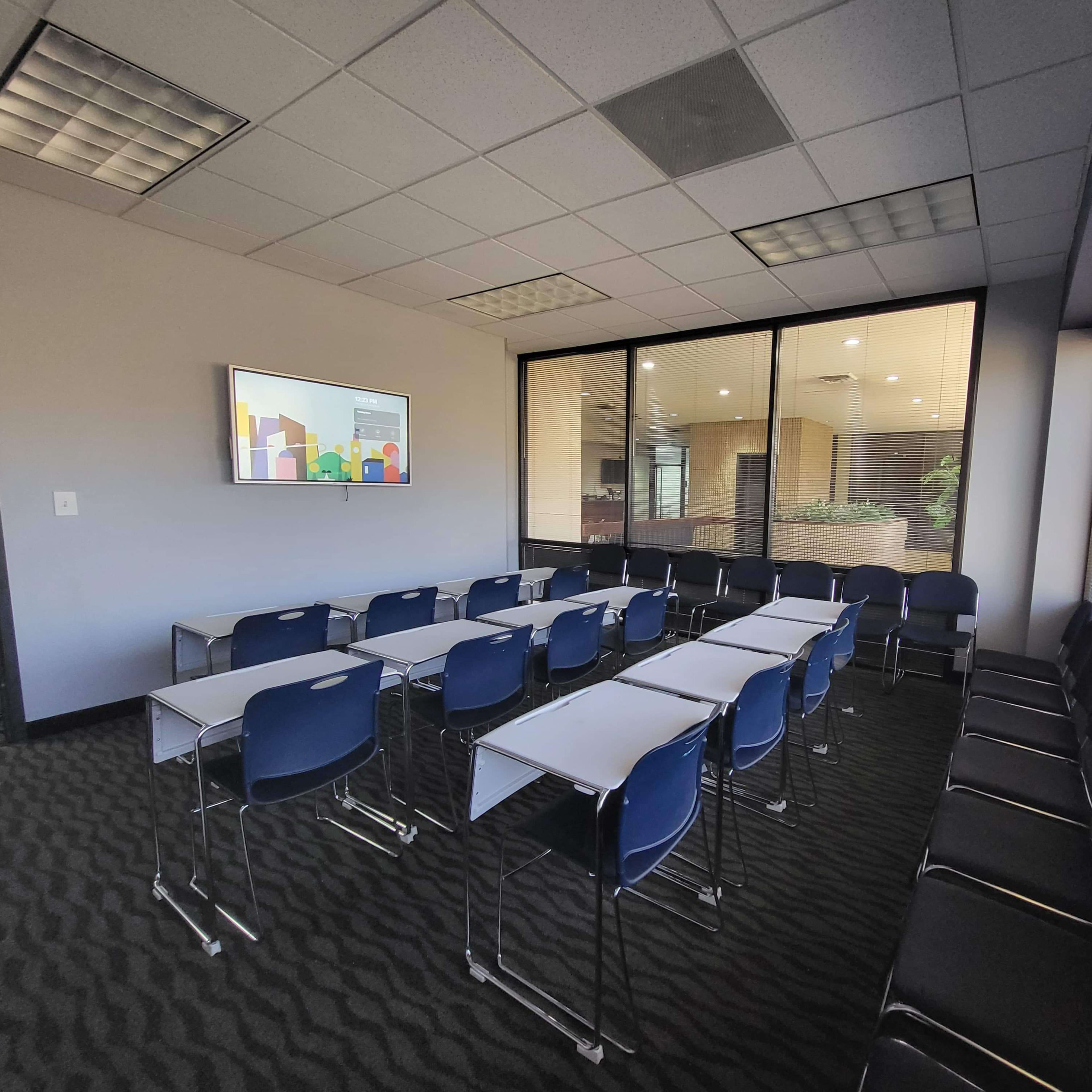 A classroom-style meeting room with rows of white tables and blue chairs, equipped with a wall-mounted screen.