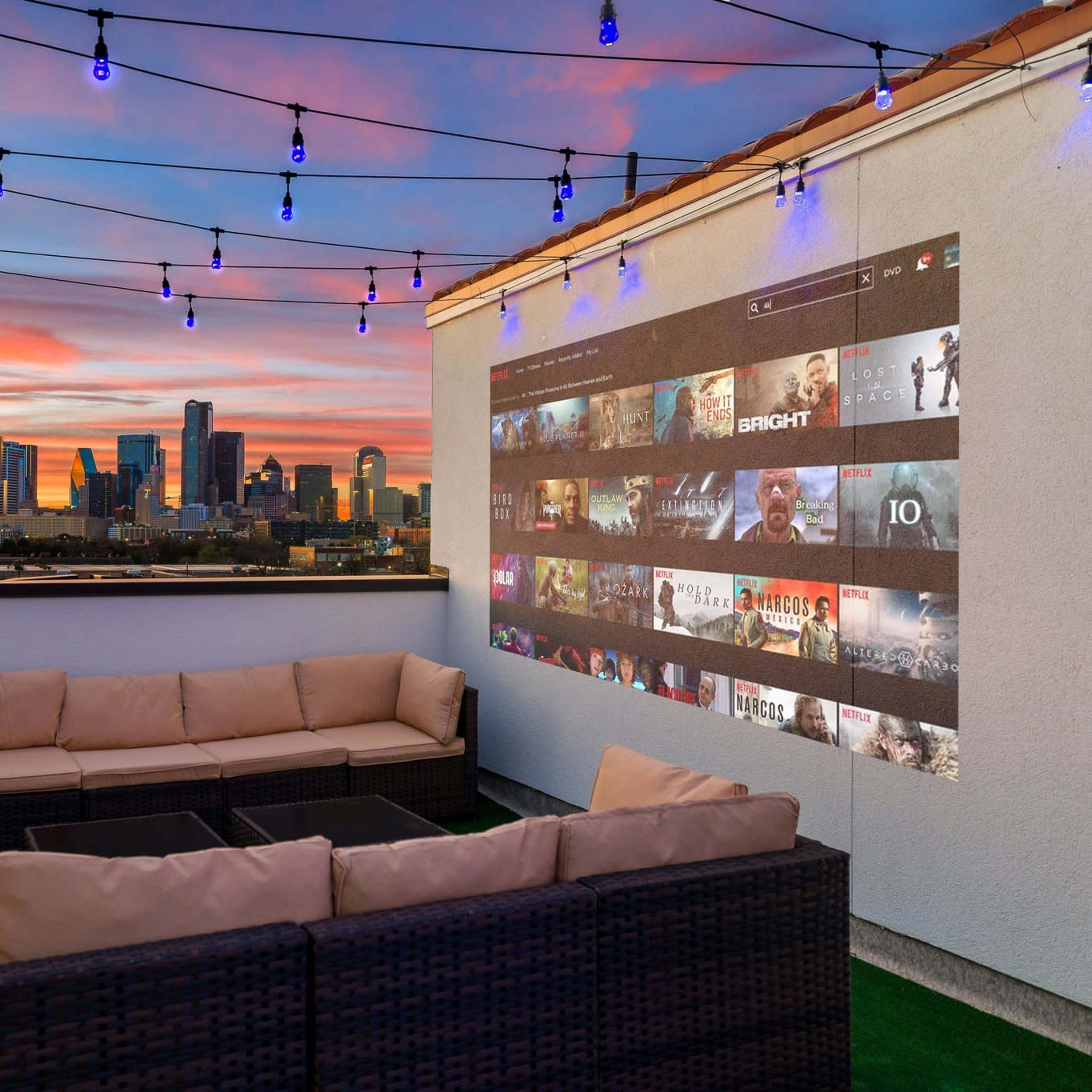 A rooftop lounge features a sectional sofa and a movie projection against a city skyline at sunset, illuminated by string lights.