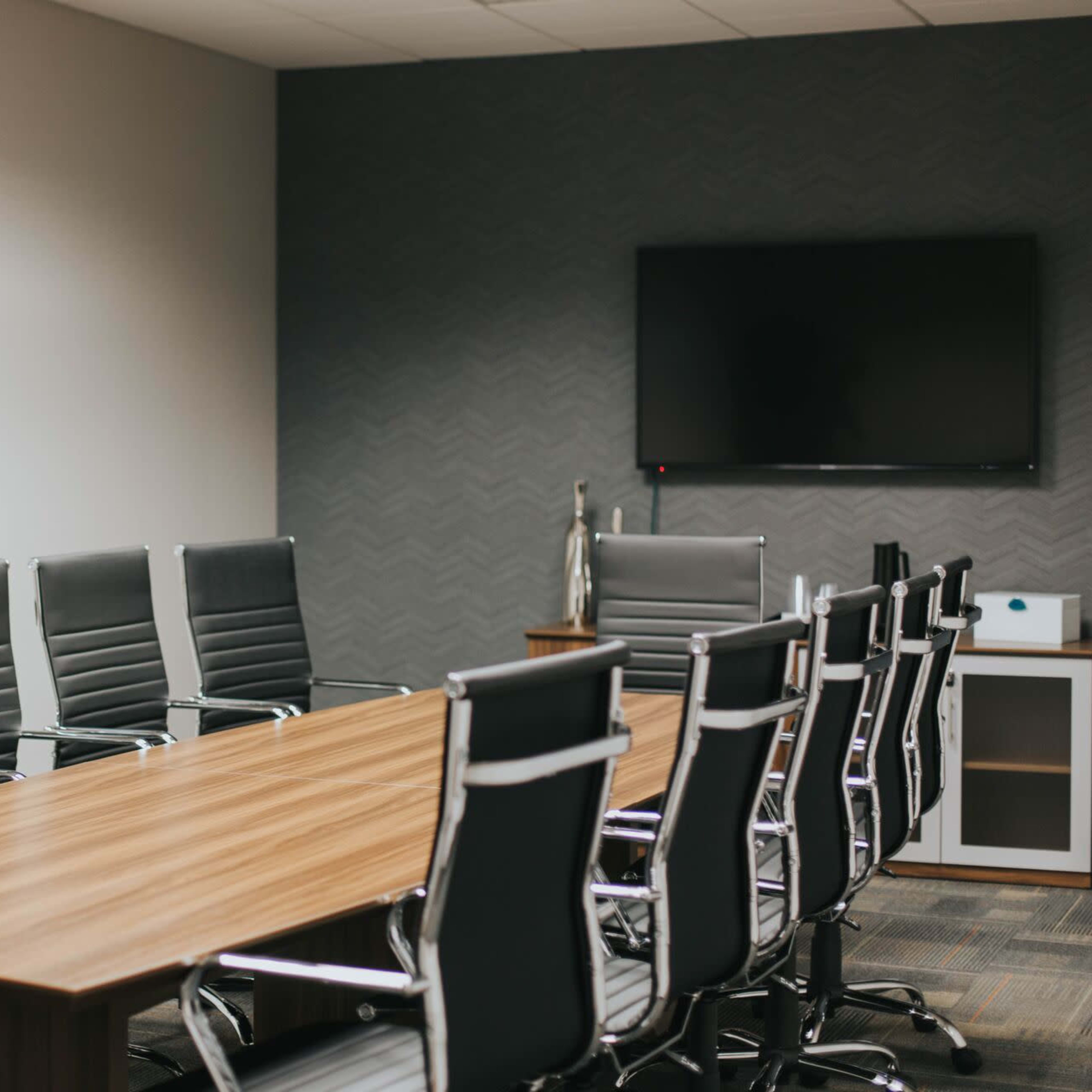 A modern conference room features a wooden table surrounded by executive-style leather chairs and a wall-mounted television.