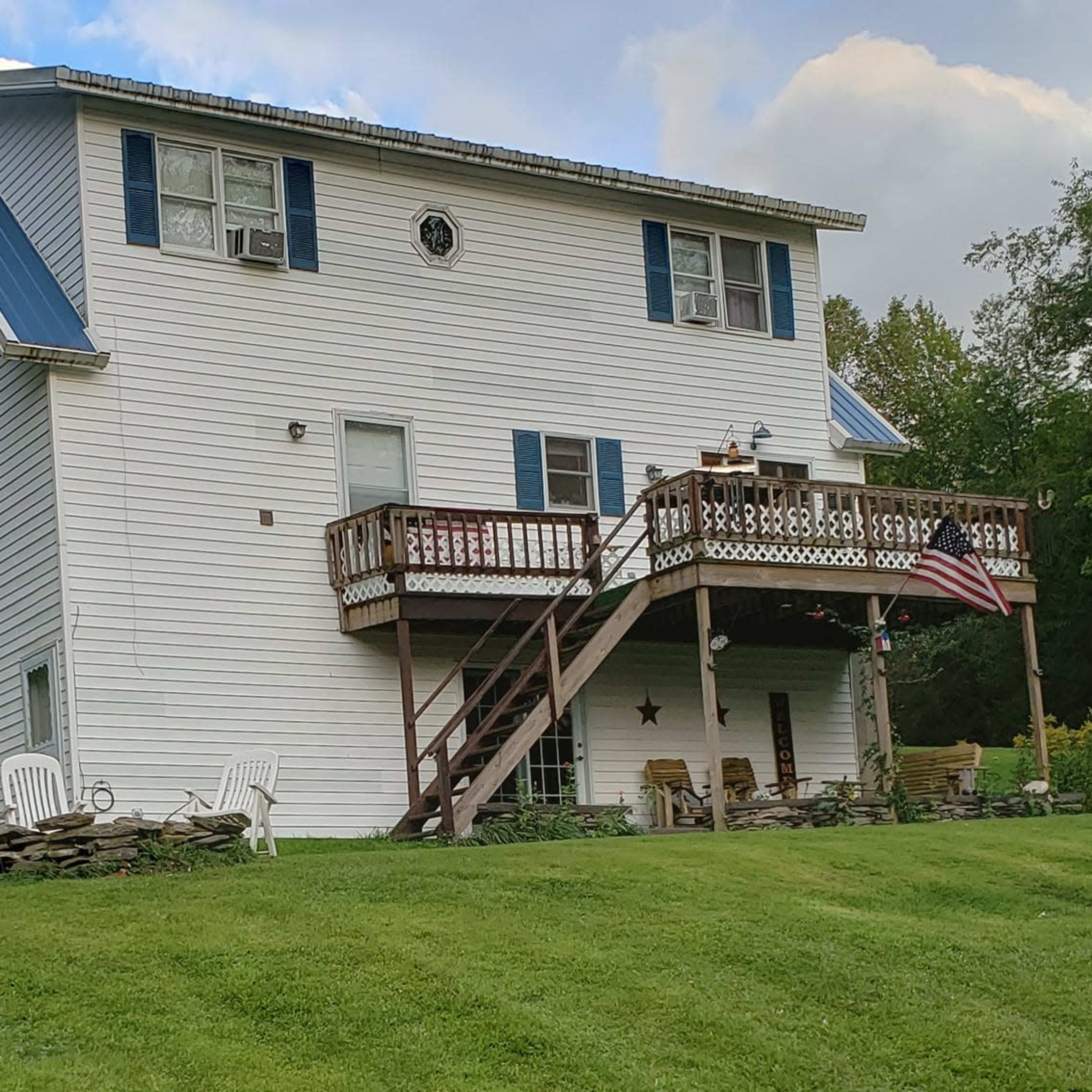 A two-story white house with a blue metal roof features a front deck and an American flag, surrounded by a grassy lawn and several white chairs.