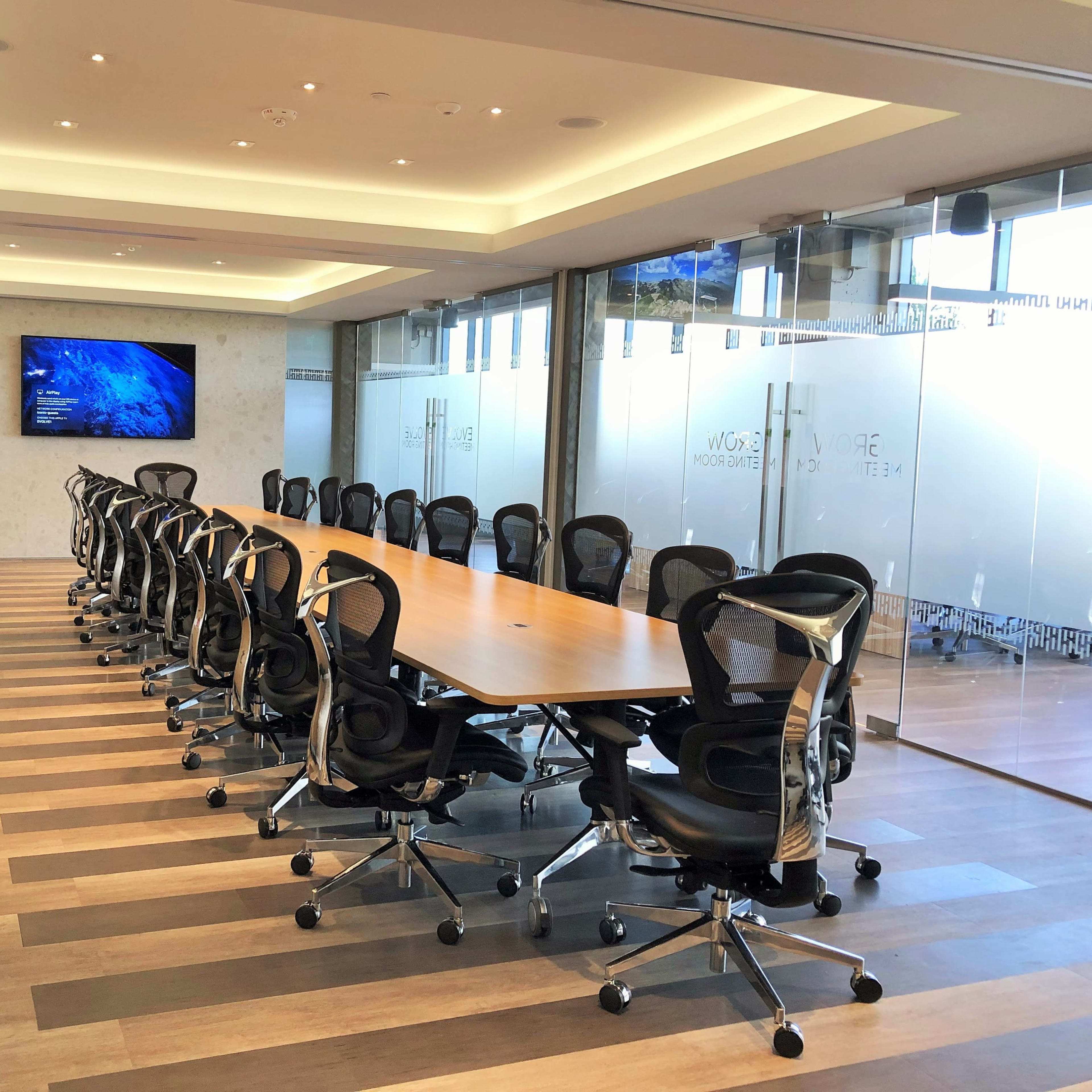 A large conference room features a long wooden table surrounded by ergonomic chairs and equipped with a wall-mounted screen.