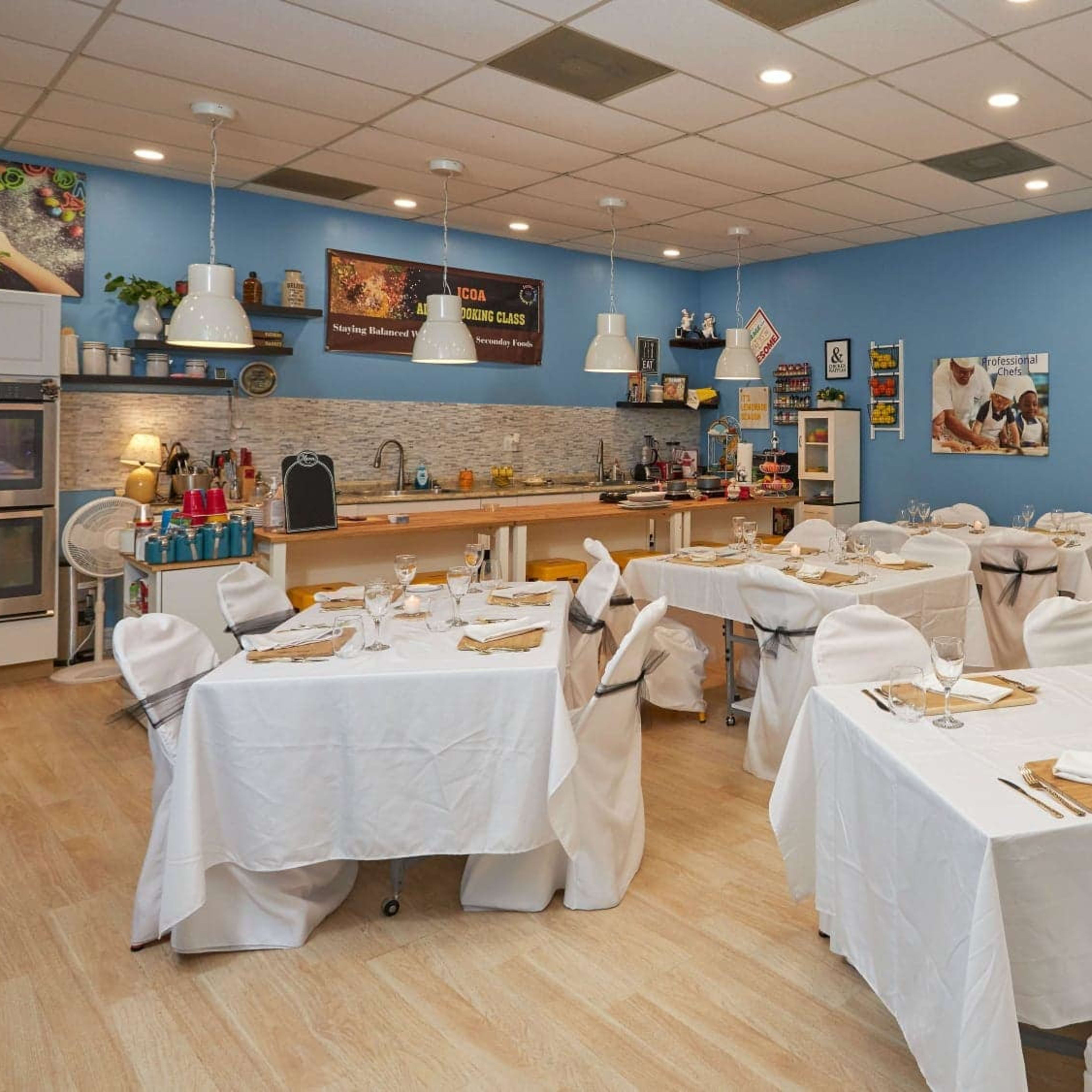 A dining area with multiple tables covered in white tablecloths, set for meals, and decorated with light blue walls and a kitchen counter in the background.