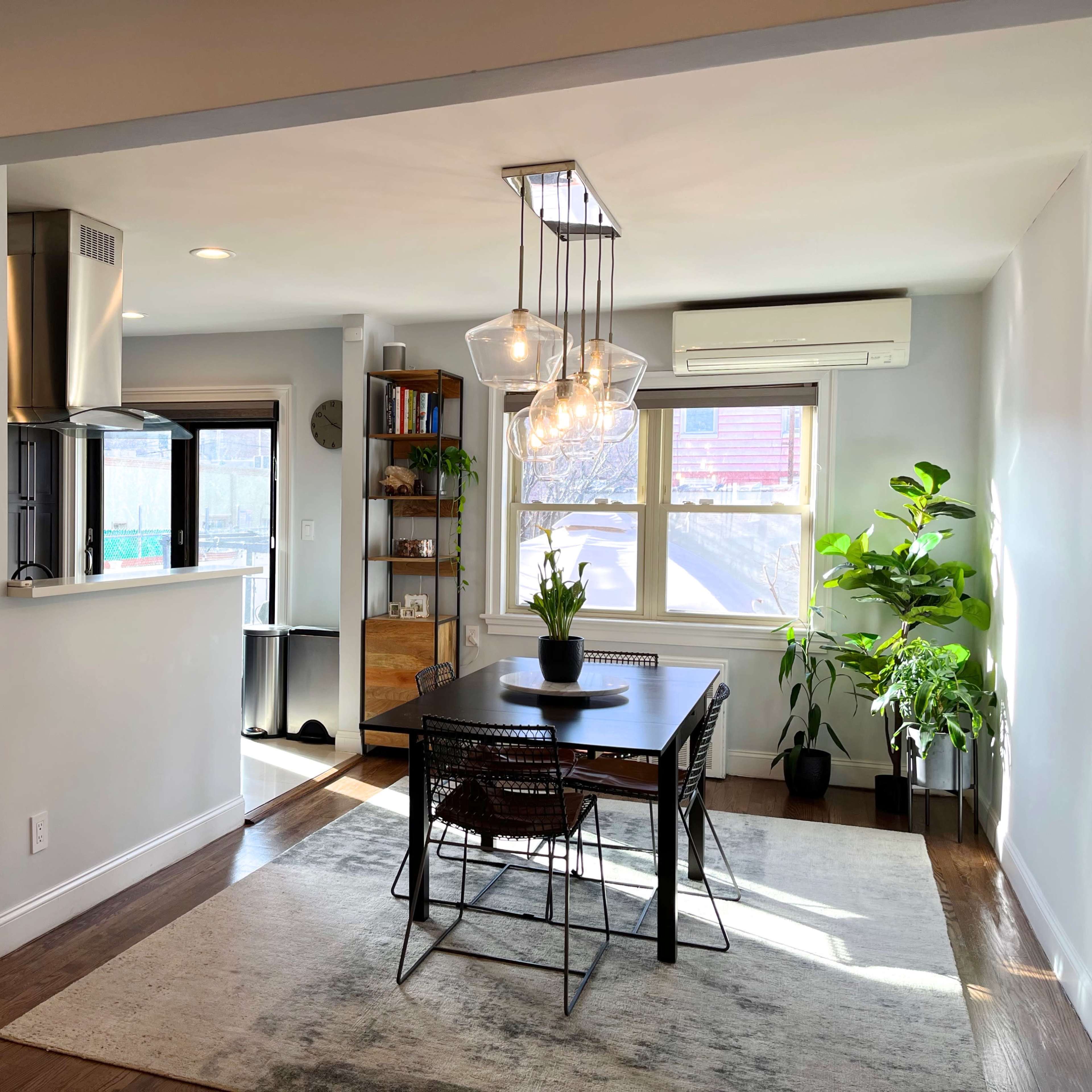 A modern dining area with a black table surrounded by chairs, illuminated by pendant lights, and featuring plants, a bookshelf, and large windows.