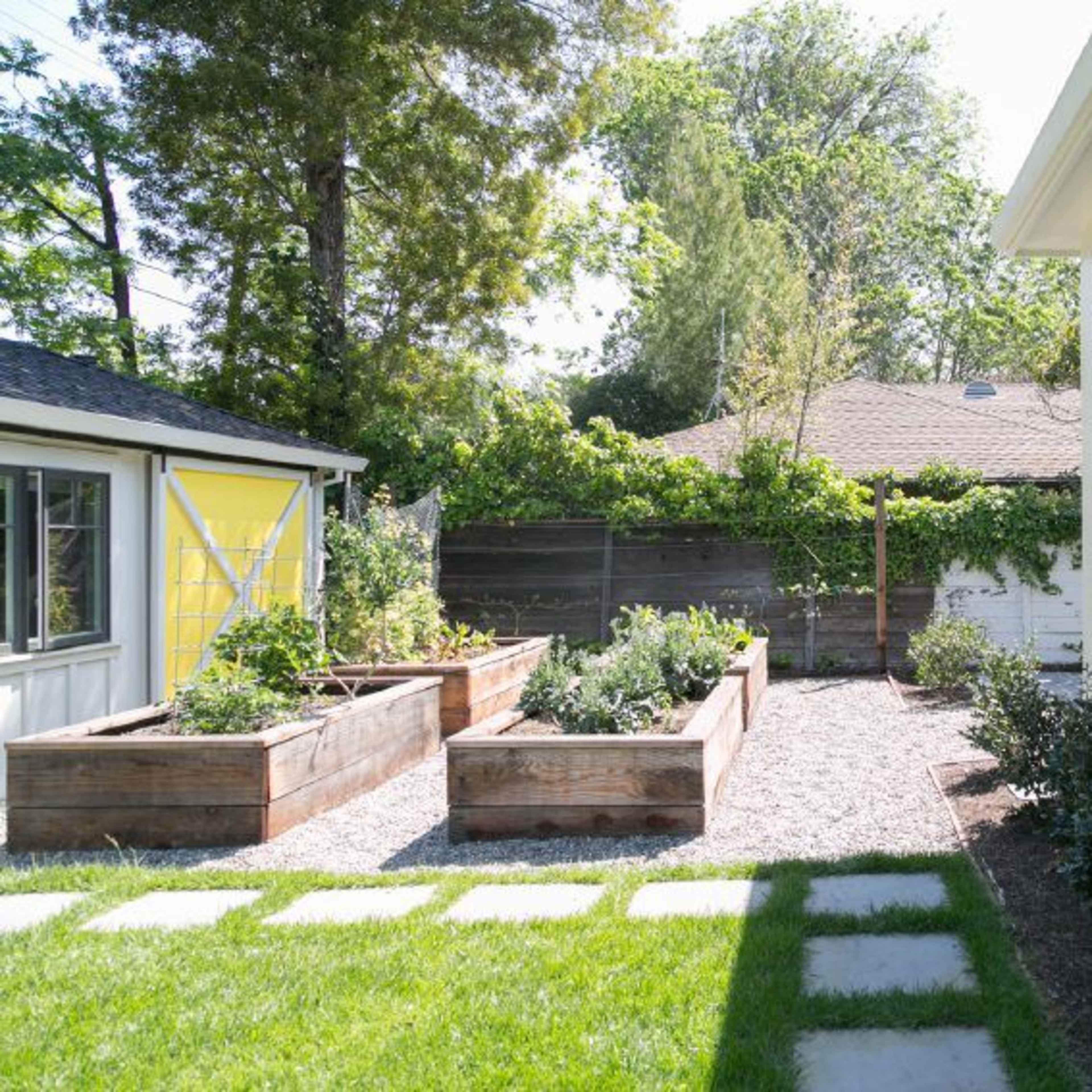 A backyard featuring raised garden beds filled with plants, a gravel pathway, and a porch area attached to a light-colored house.
