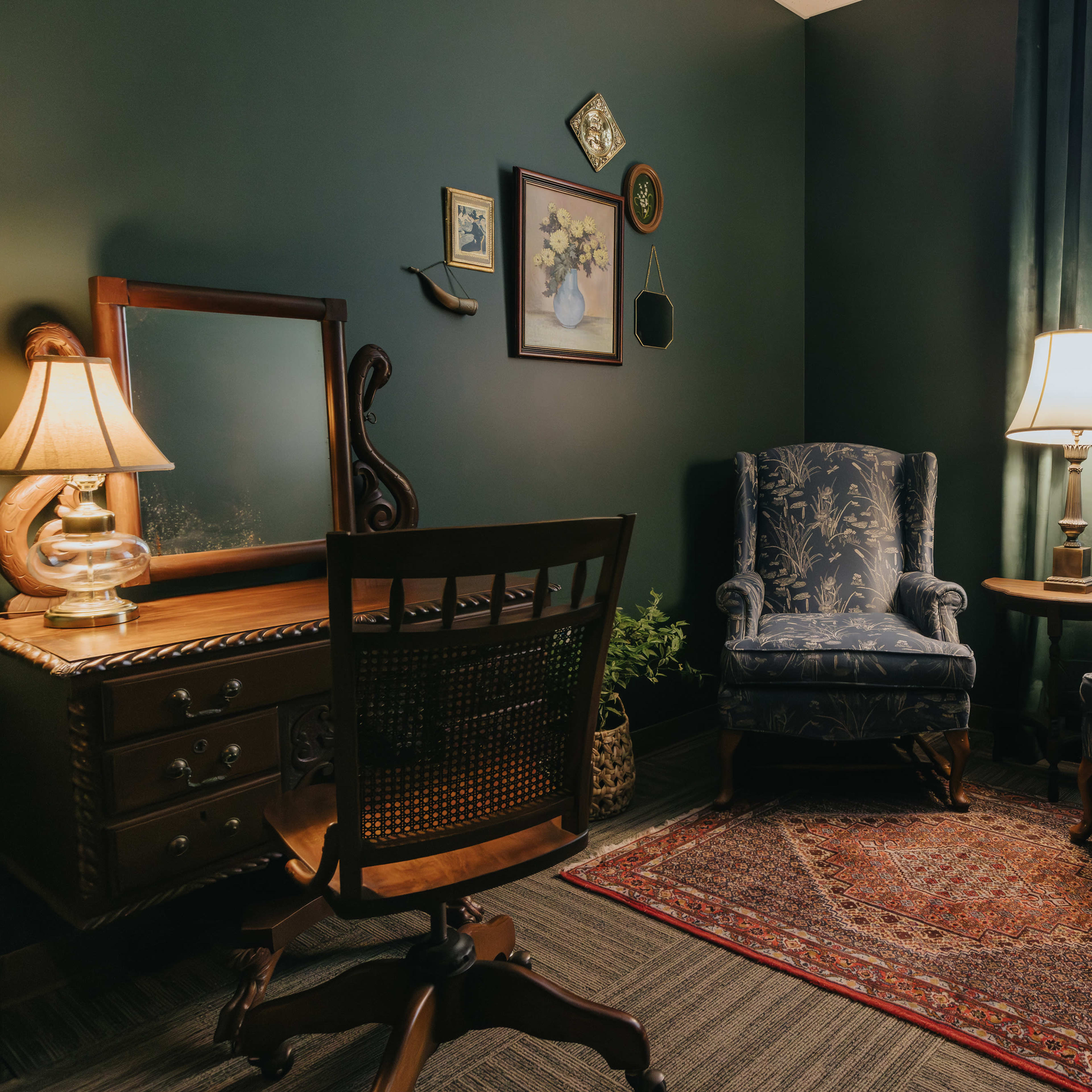 A cozy corner of a room featuring two upholstered chairs, a wooden desk with a mirror, a small side table with a lamp, and a patterned rug.