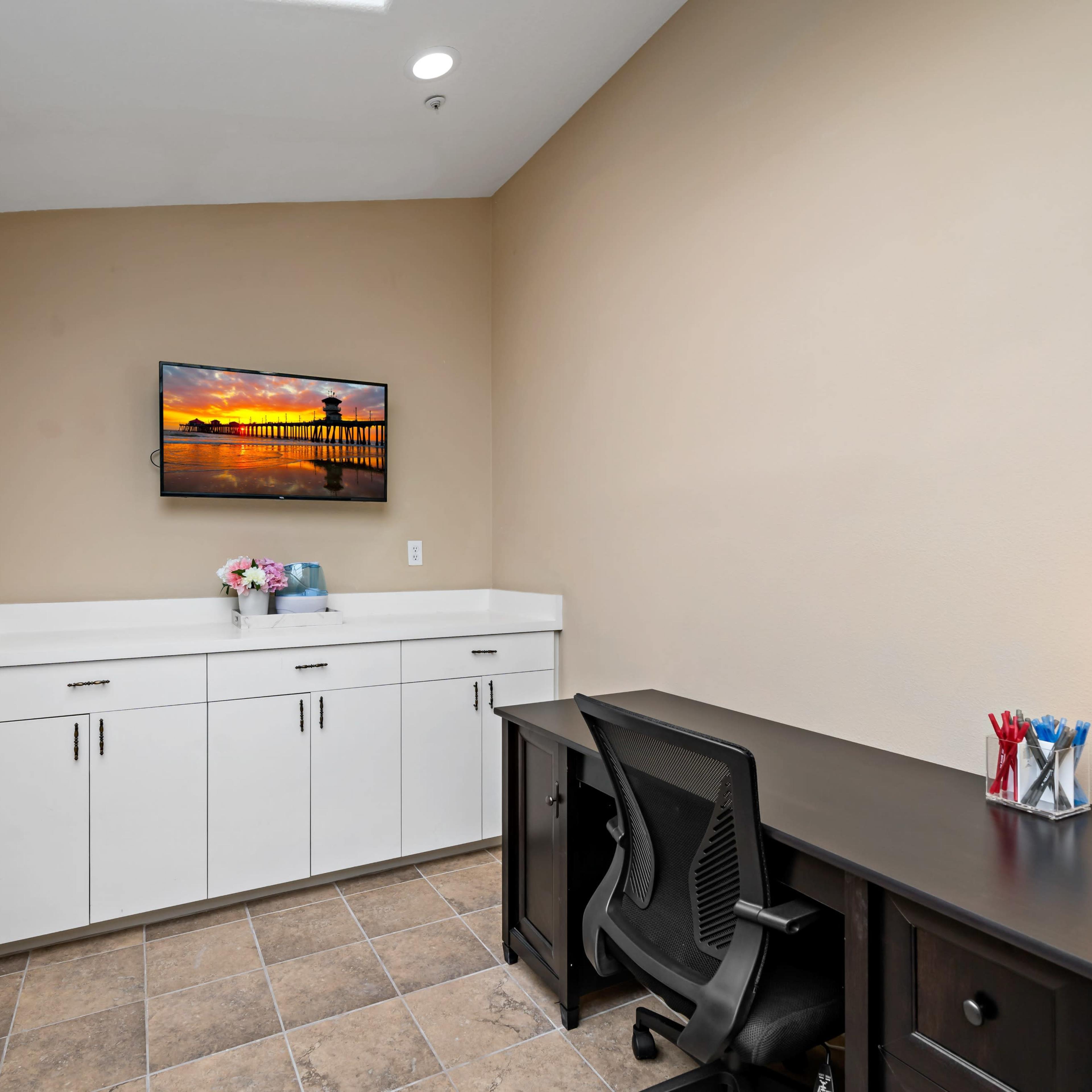 A small office space features a black desk with a chair, white cabinetry, and a mounted television displaying a sunset over a pier.