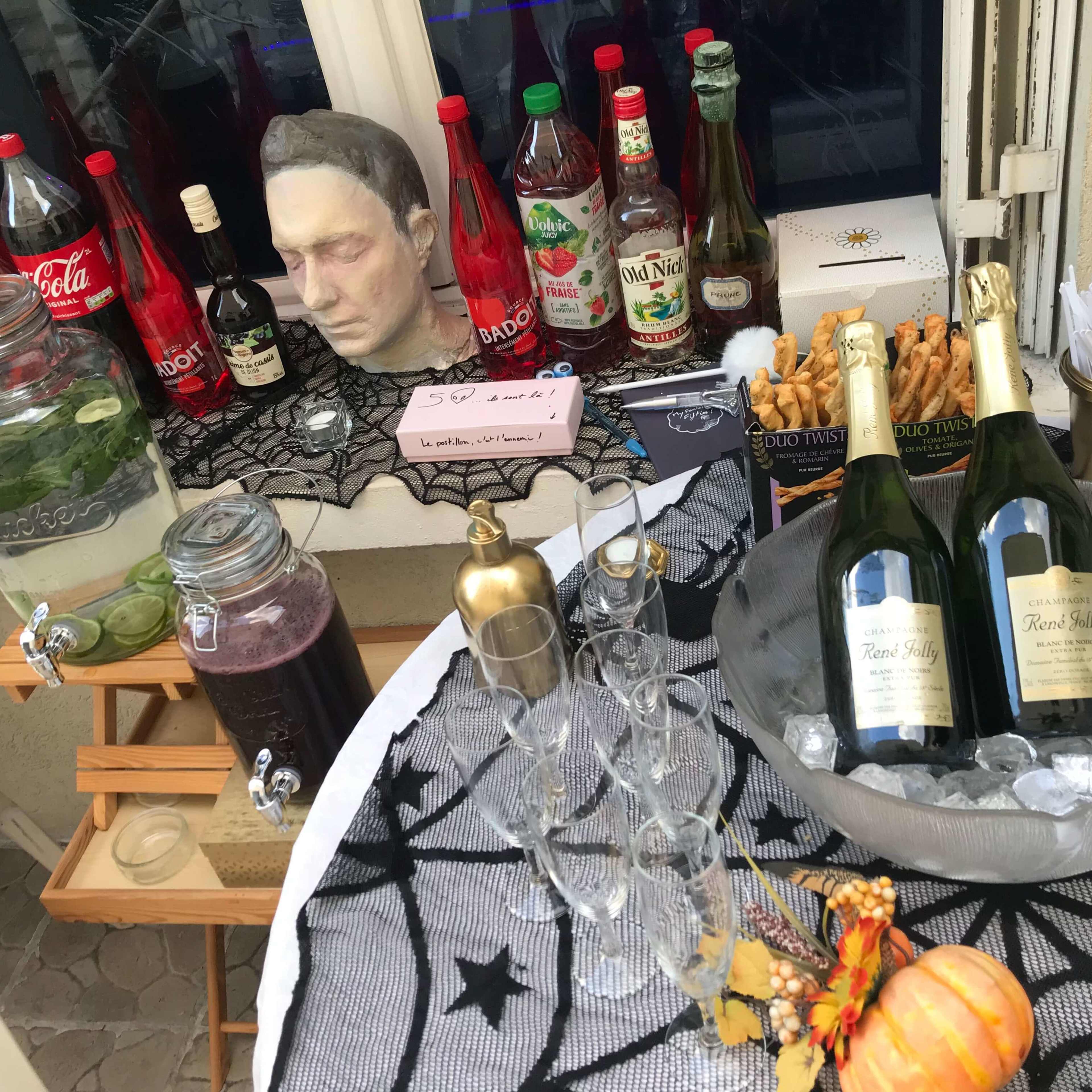 A table is set with various beverages, snacks, and decorations, including bottles of soda, a head sculpture, and sparkling wine on ice.