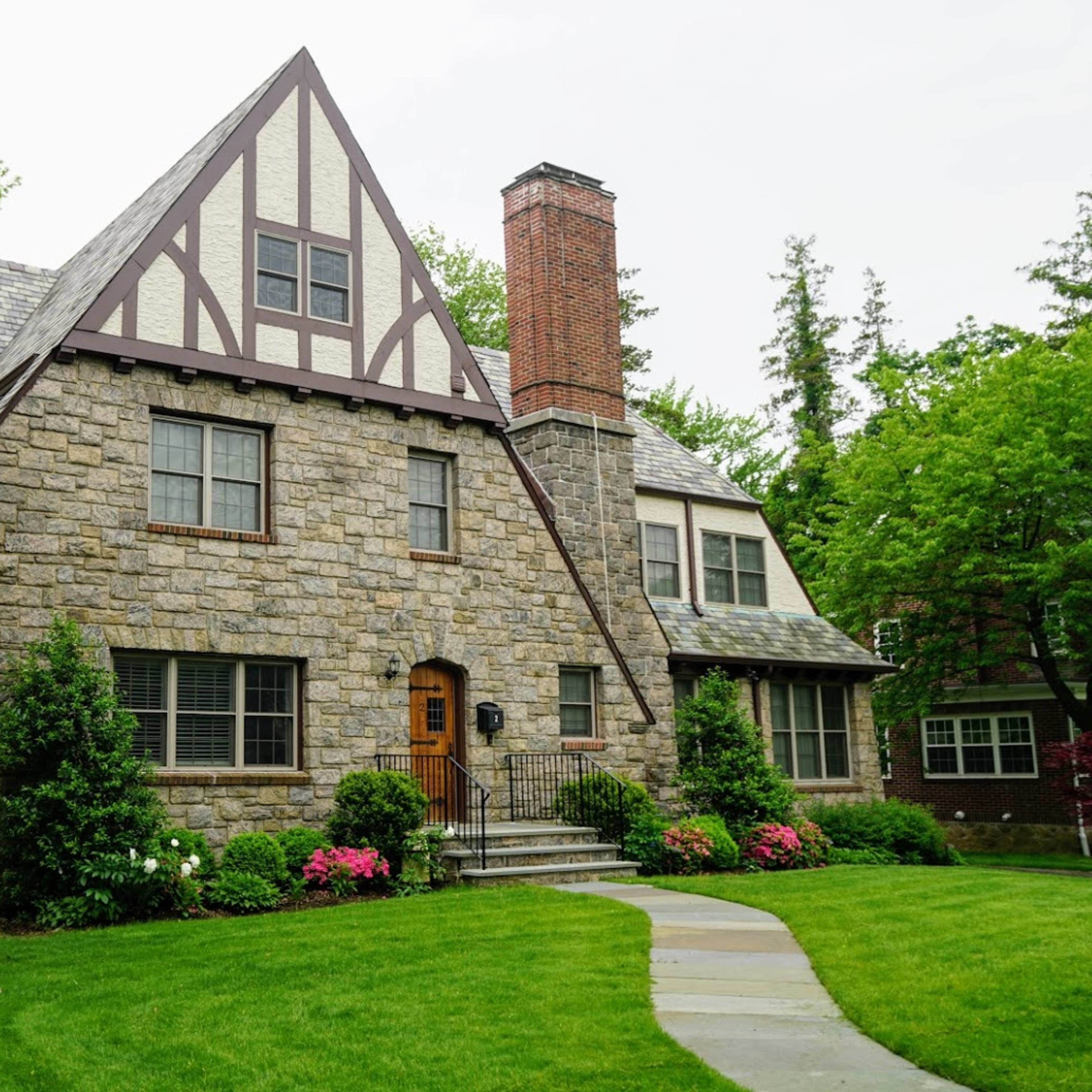 A stone house with a steeply pitched roof and a central wooden door is surrounded by a well-maintained lawn and shrubs.
