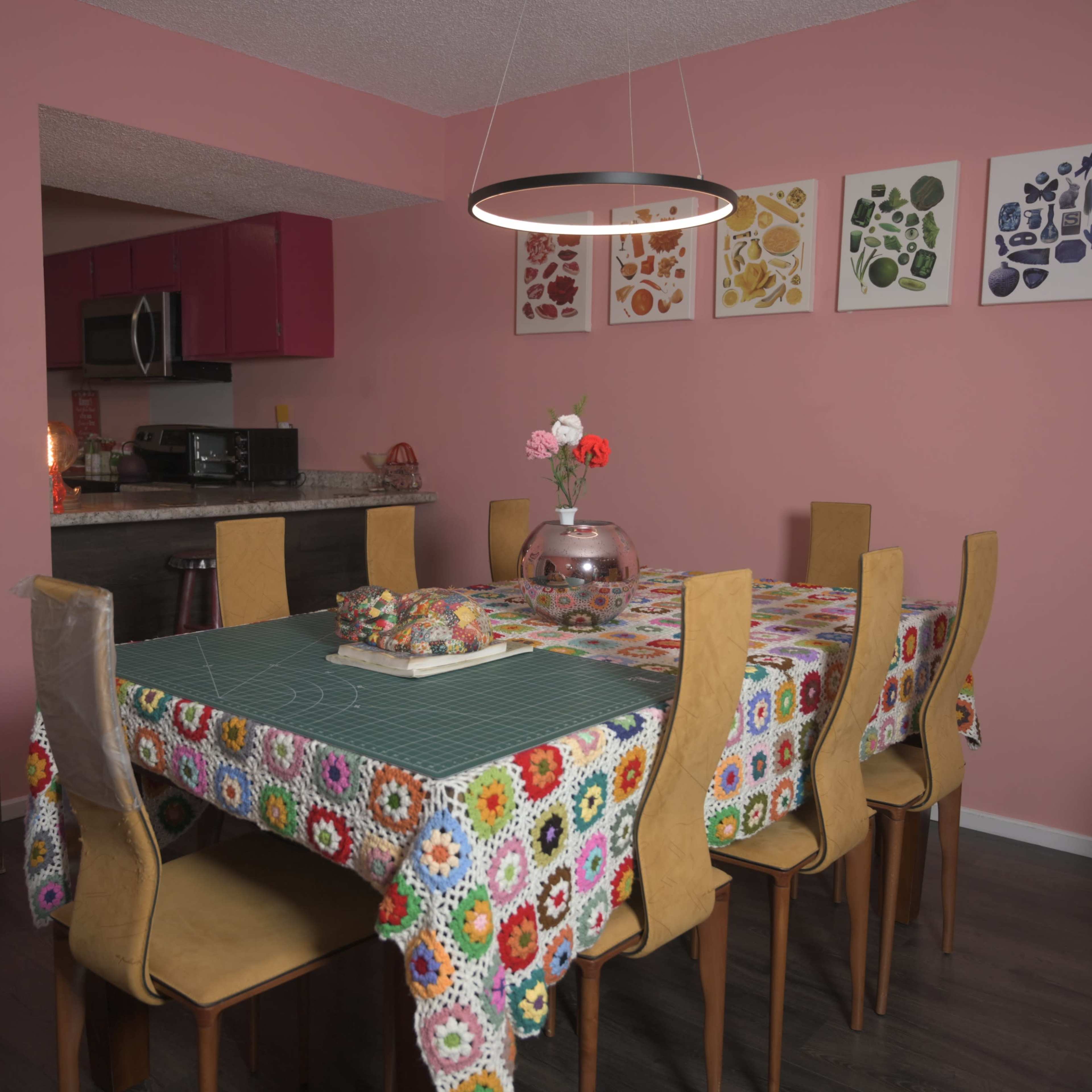 A dining room features a large table with a colorful tablecloth, surrounded by wooden chairs, and walls painted pink adorned with framed art pieces.