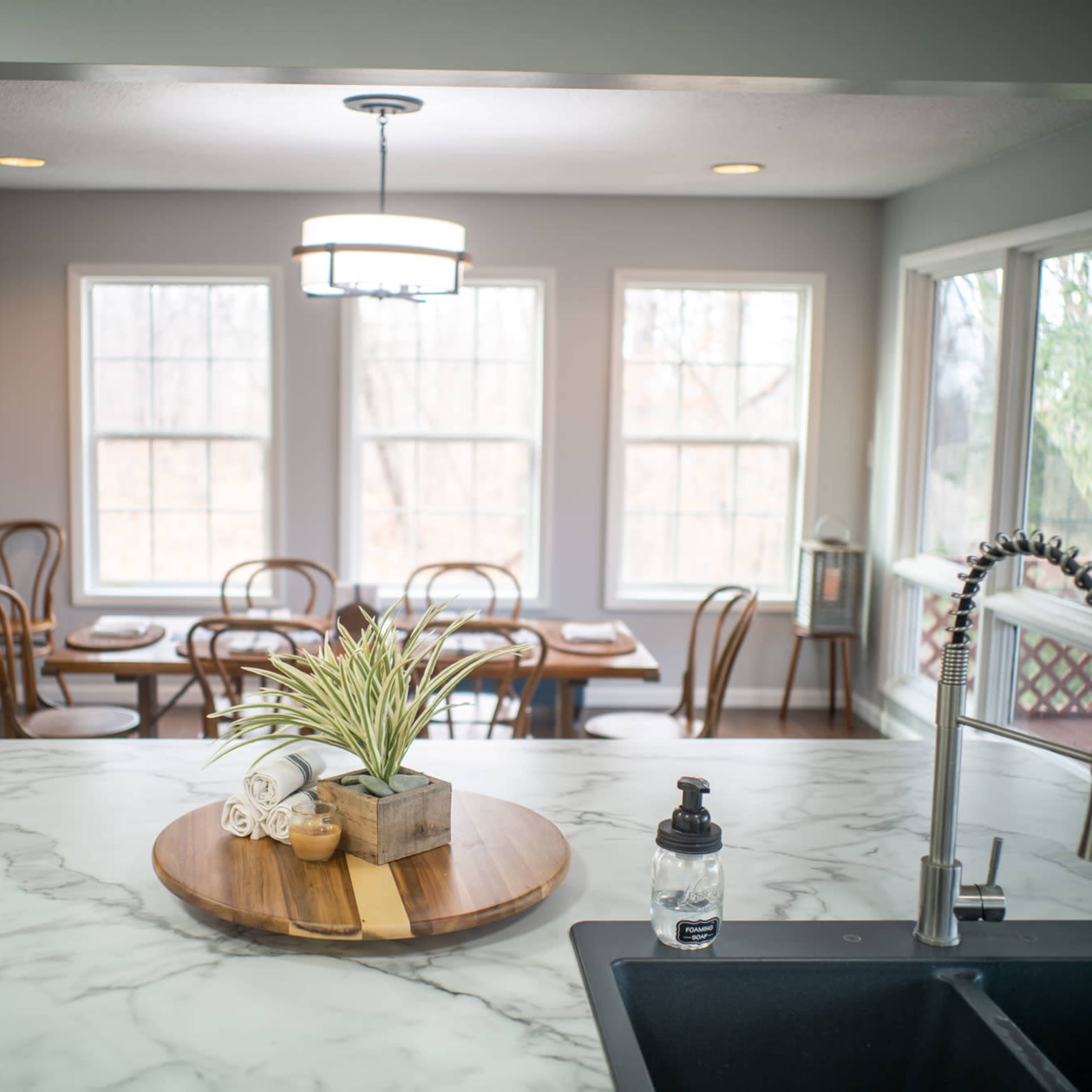A dining area features several chairs around a table, with large windows providing natural light, and a kitchen island with a marble countertop in the foreground.