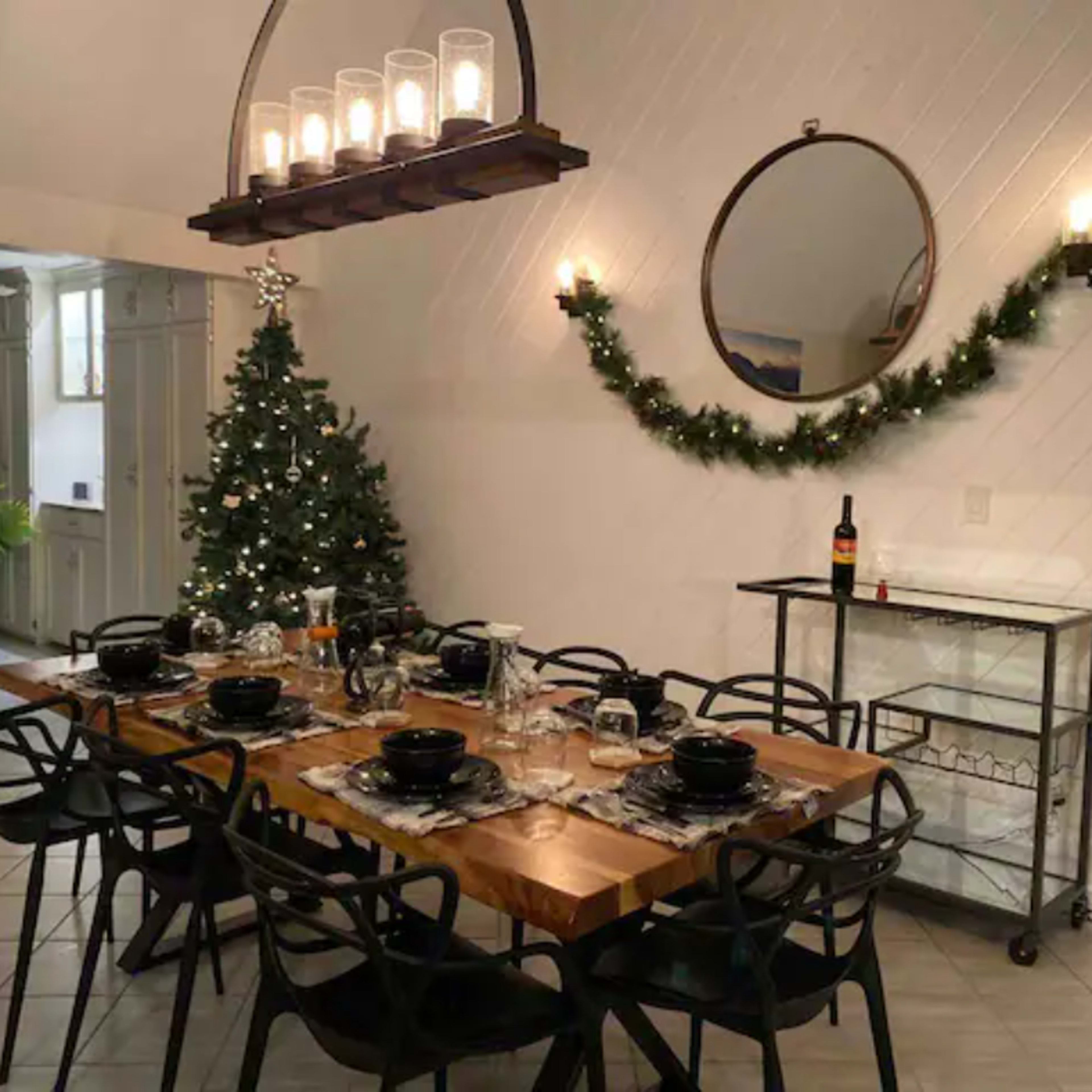 A dining area with a long wooden table set for a meal, surrounded by black chairs, a Christmas tree in the corner, and decorative lighting overhead.