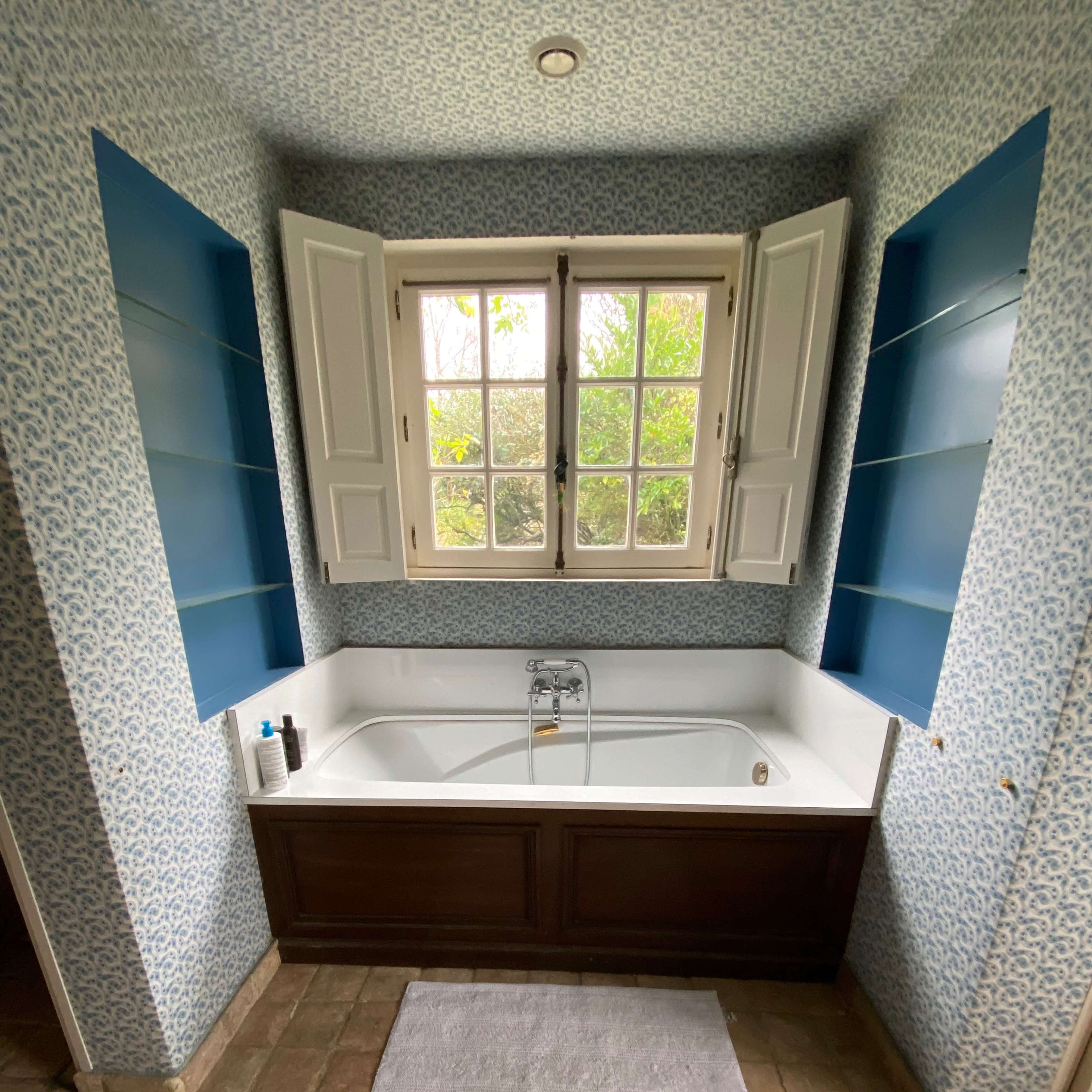 A bathroom with a rectangular bathtub positioned beneath a window, surrounded by patterned wallpaper and blue shelving.