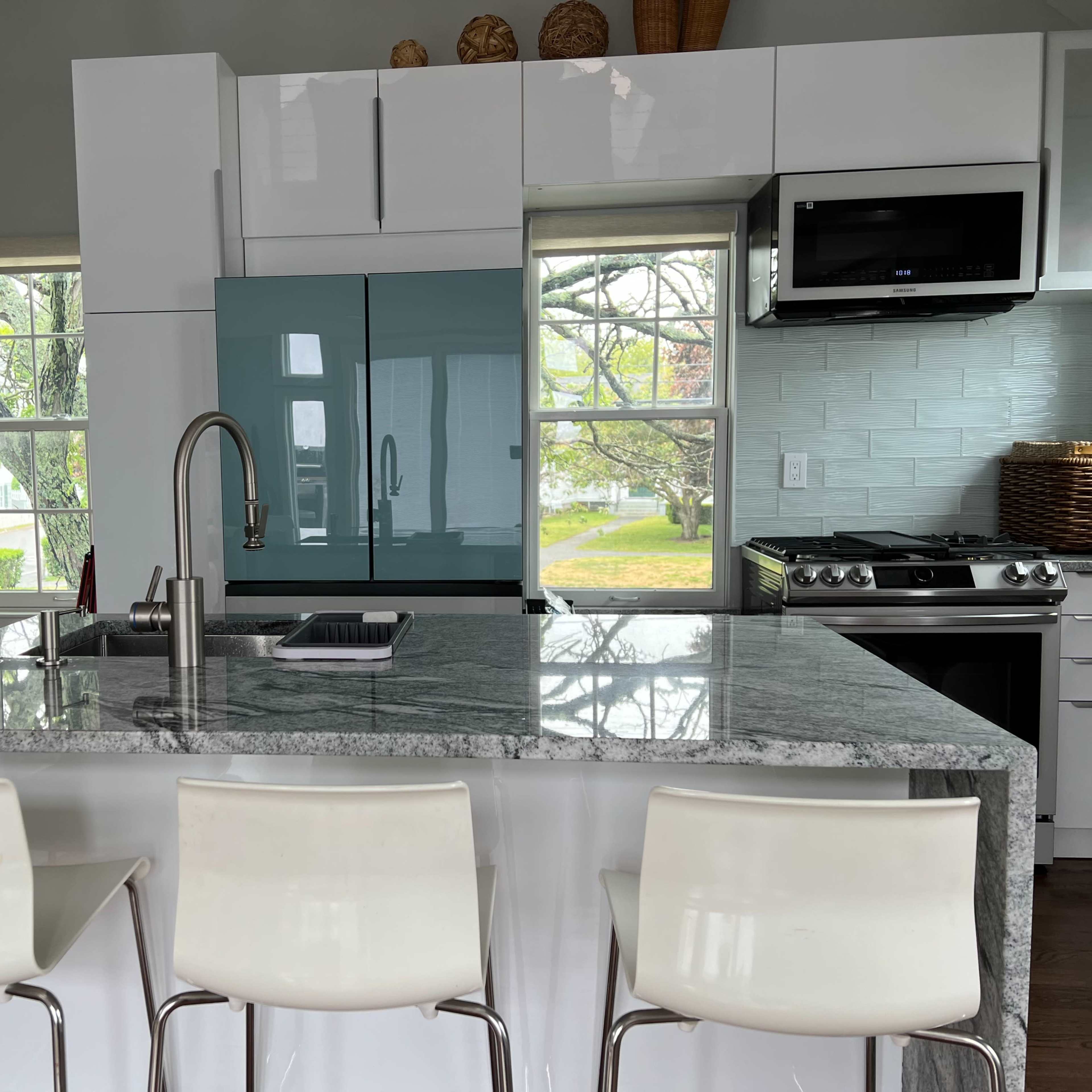 A modern kitchen features a large granite island with three white stools, a blue refrigerator, and large windows overlooking a garden.