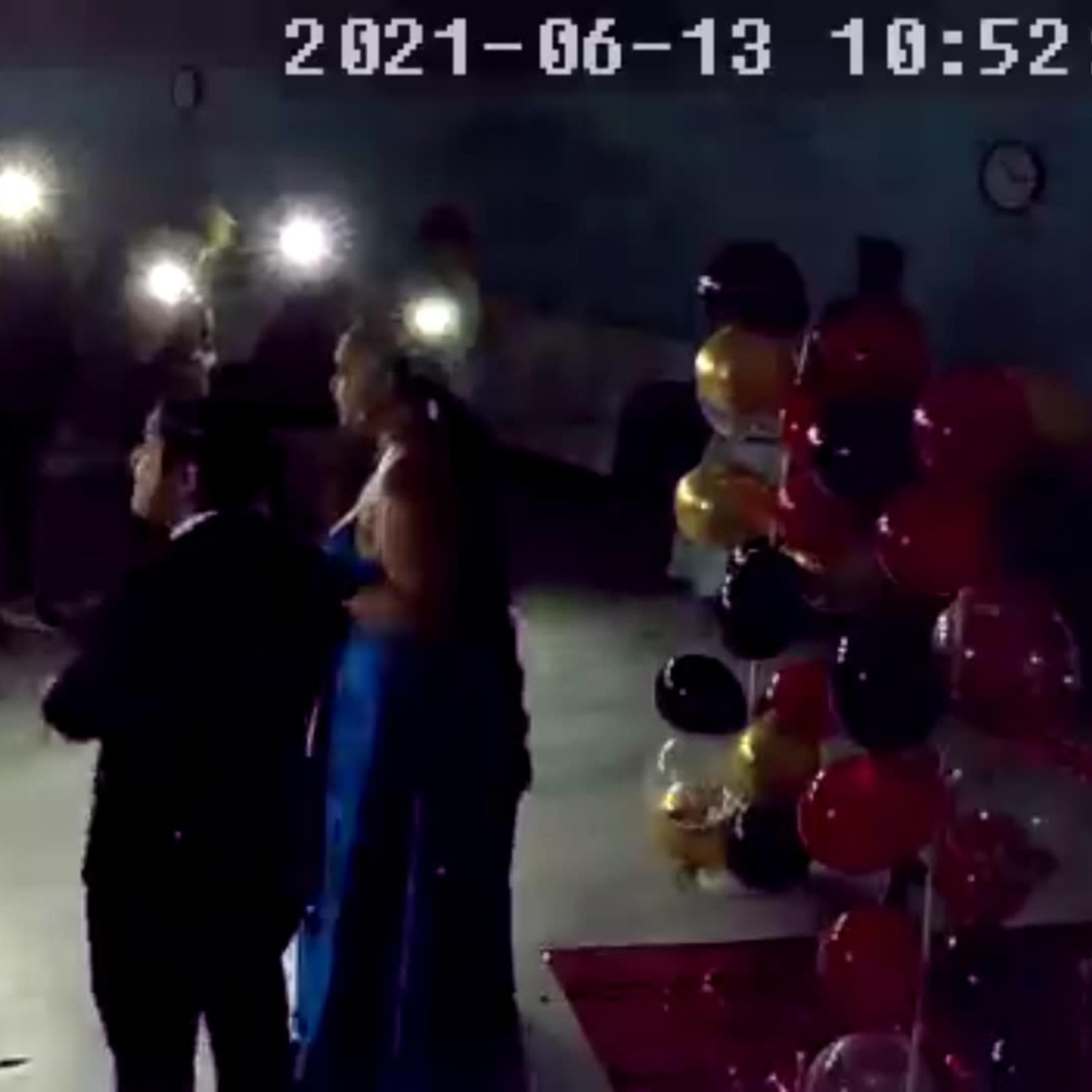A couple stands on a dance floor surrounded by party guests, some holding their phones with flashlights on, while balloons in black, red, and yellow decorate the scene.