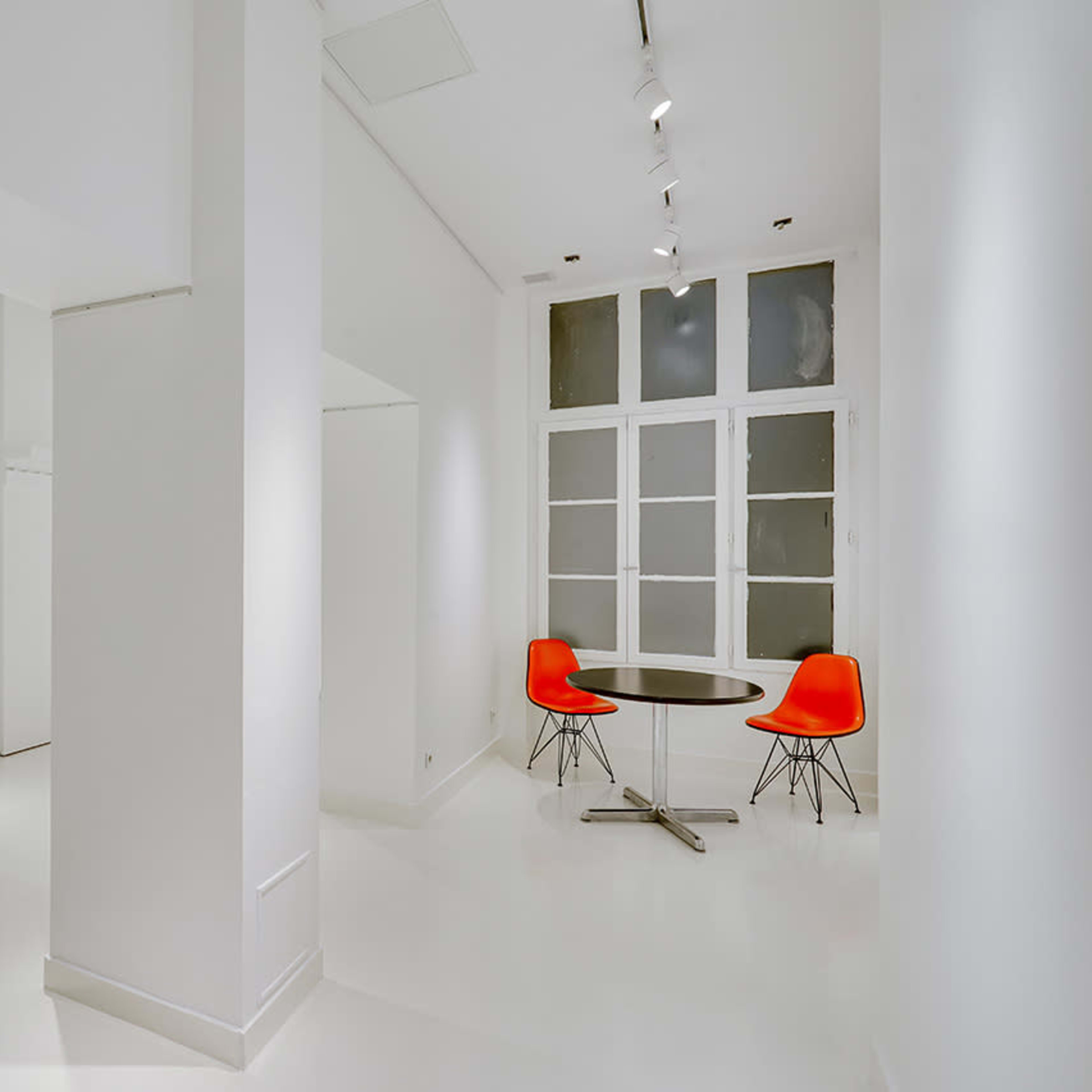 A minimalist, white-walled room featuring a round black table with two red chairs and a large window with multiple panes.
