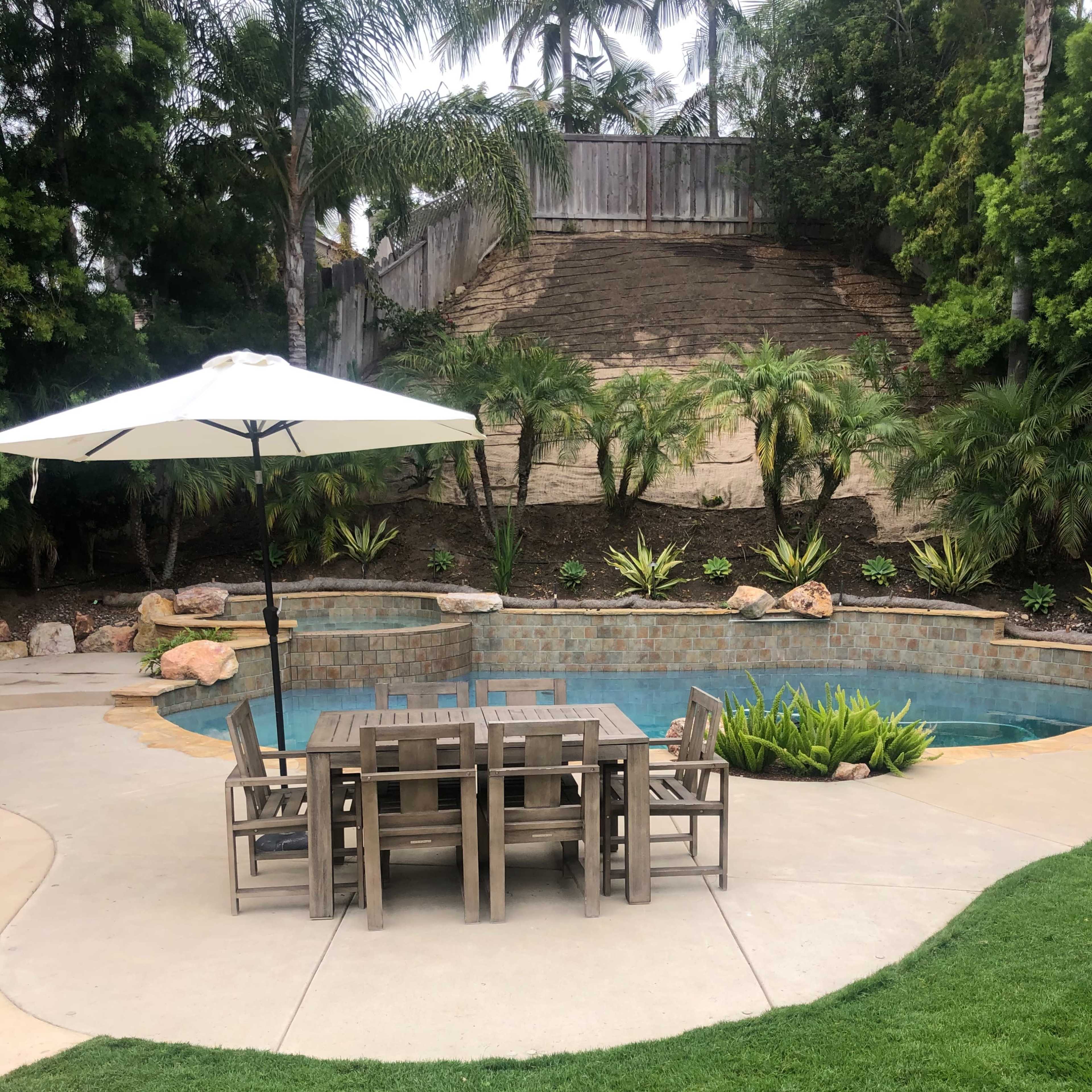 A wooden table and chairs sit under an umbrella beside a swimming pool, surrounded by lush greenery and stone steps in the background.
