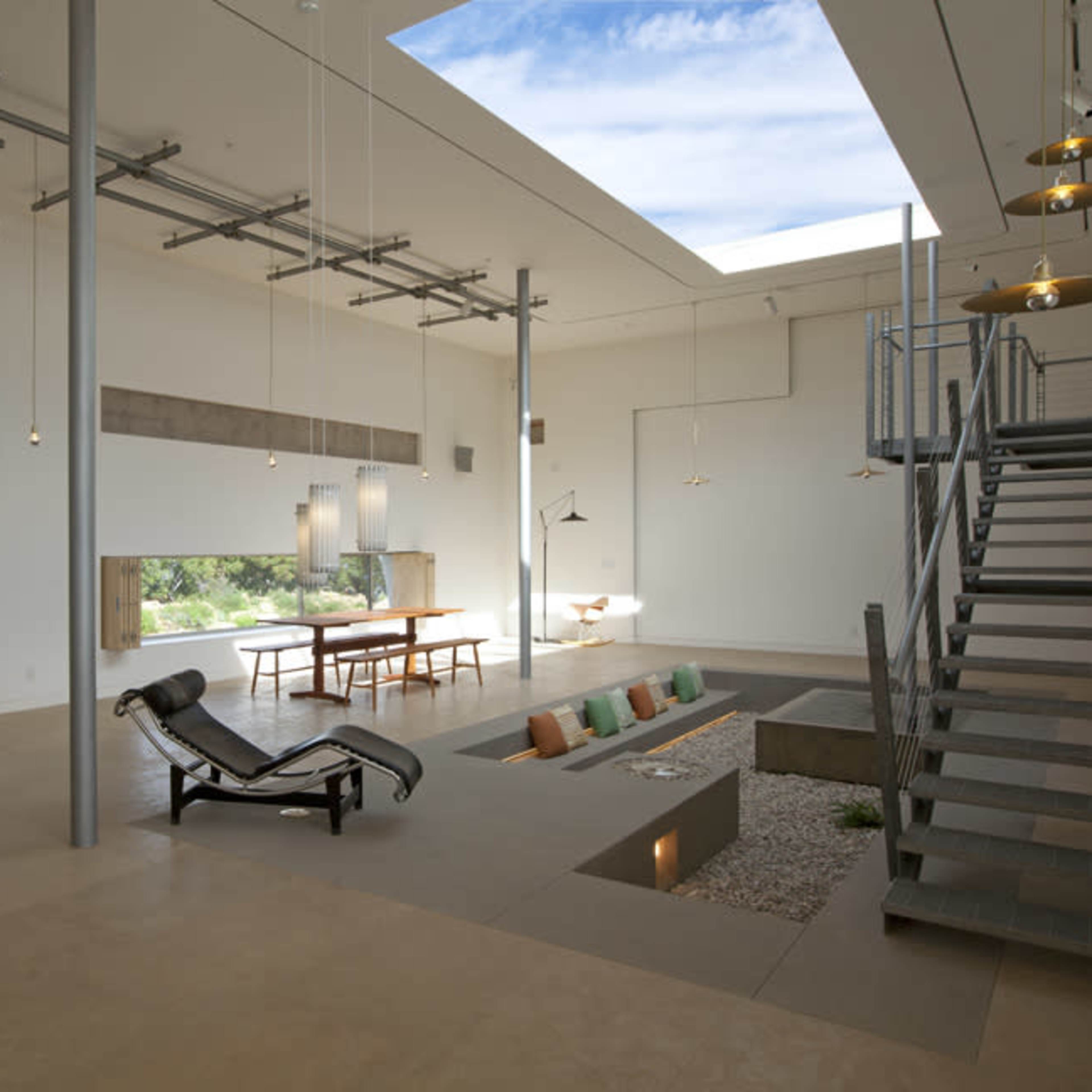 The interior of a modern, minimalistic space features a lounge area with a chaise lounge, a dining table, and large windows allowing natural light to enter.