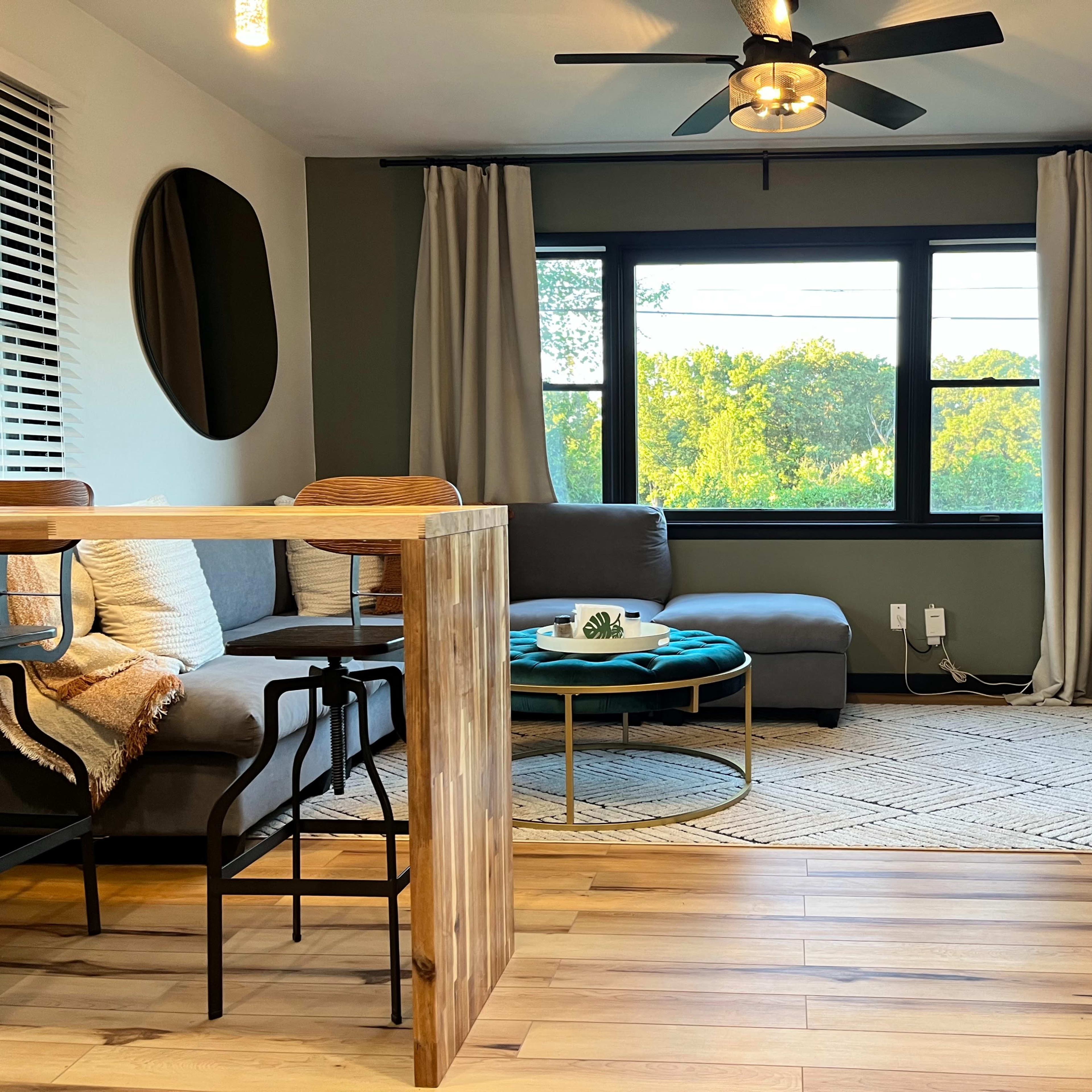 A modern living room featuring a gray sectional sofa, a round coffee table, and a large window with curtains, overlooking a green landscape.