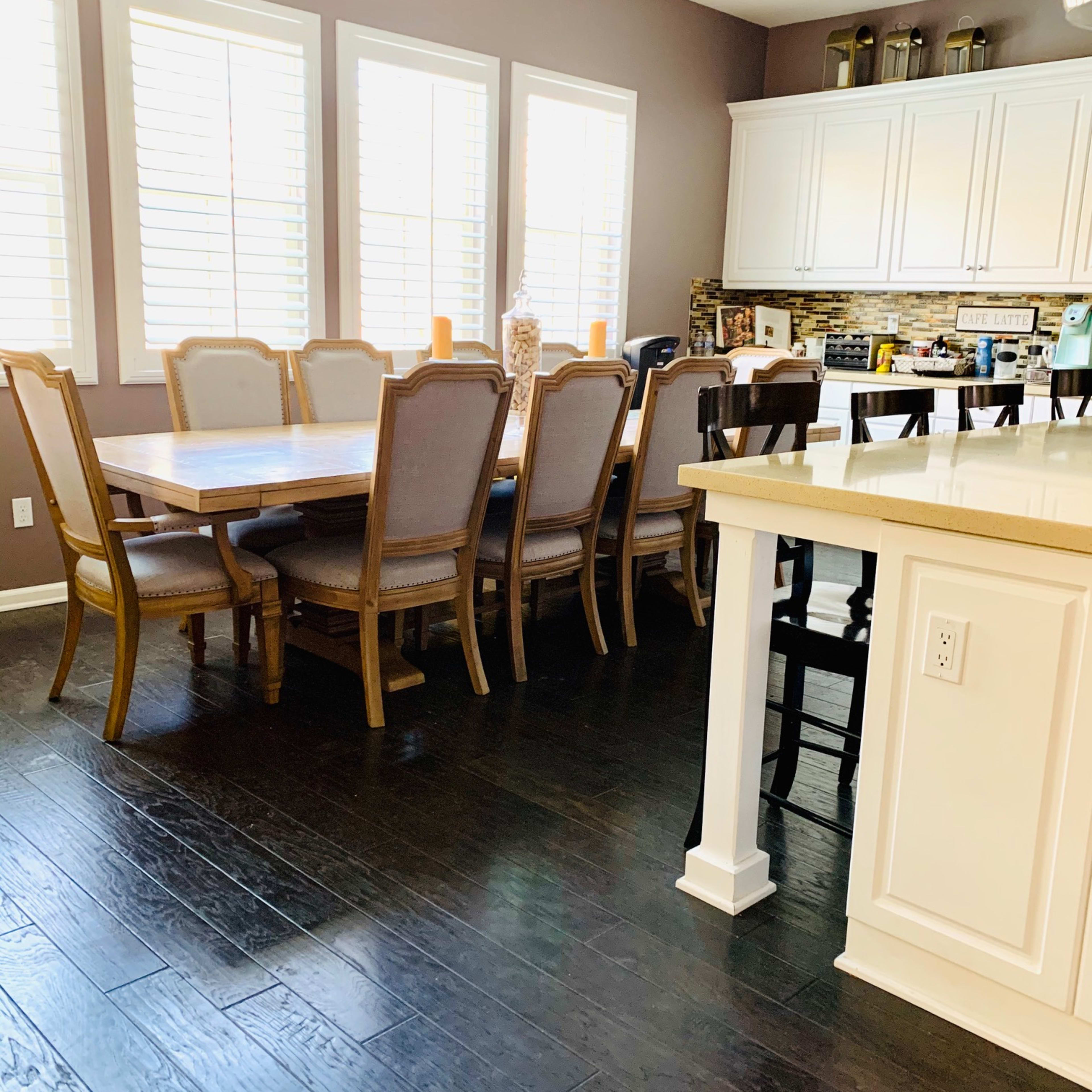 A dining area features a large wooden table surrounded by upholstered chairs, with sunlight streaming through several windows.