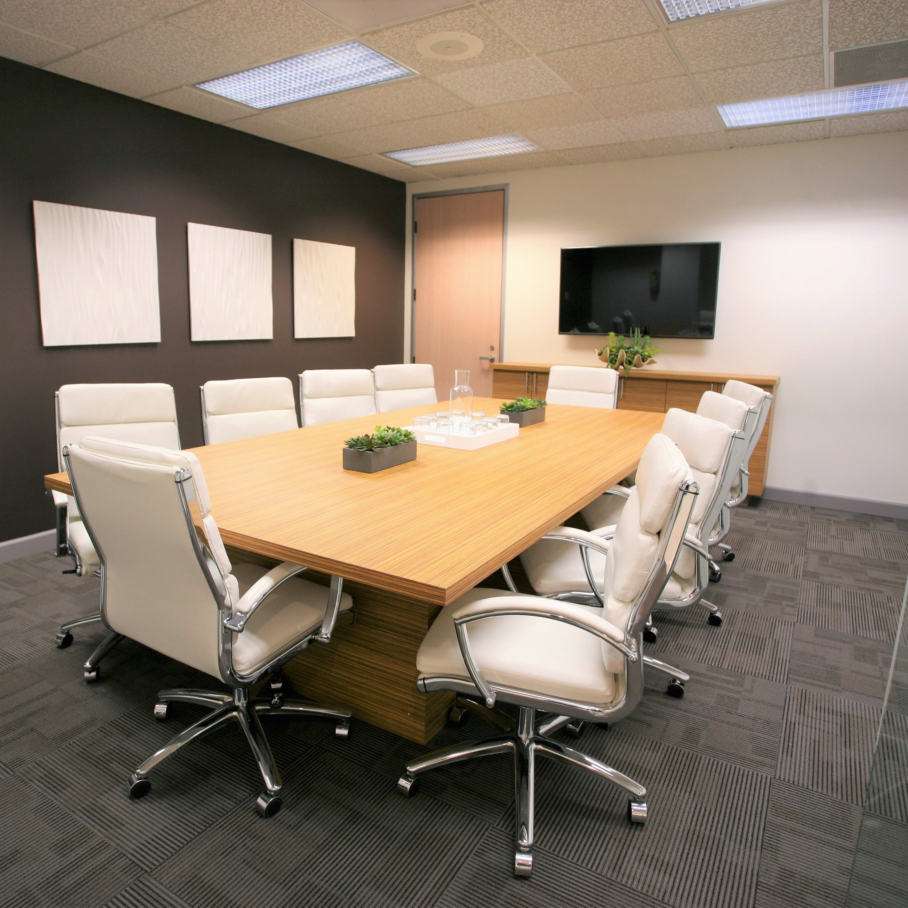 A modern conference room features a large wooden table surrounded by white chairs, with a television and potted plants on the walls.