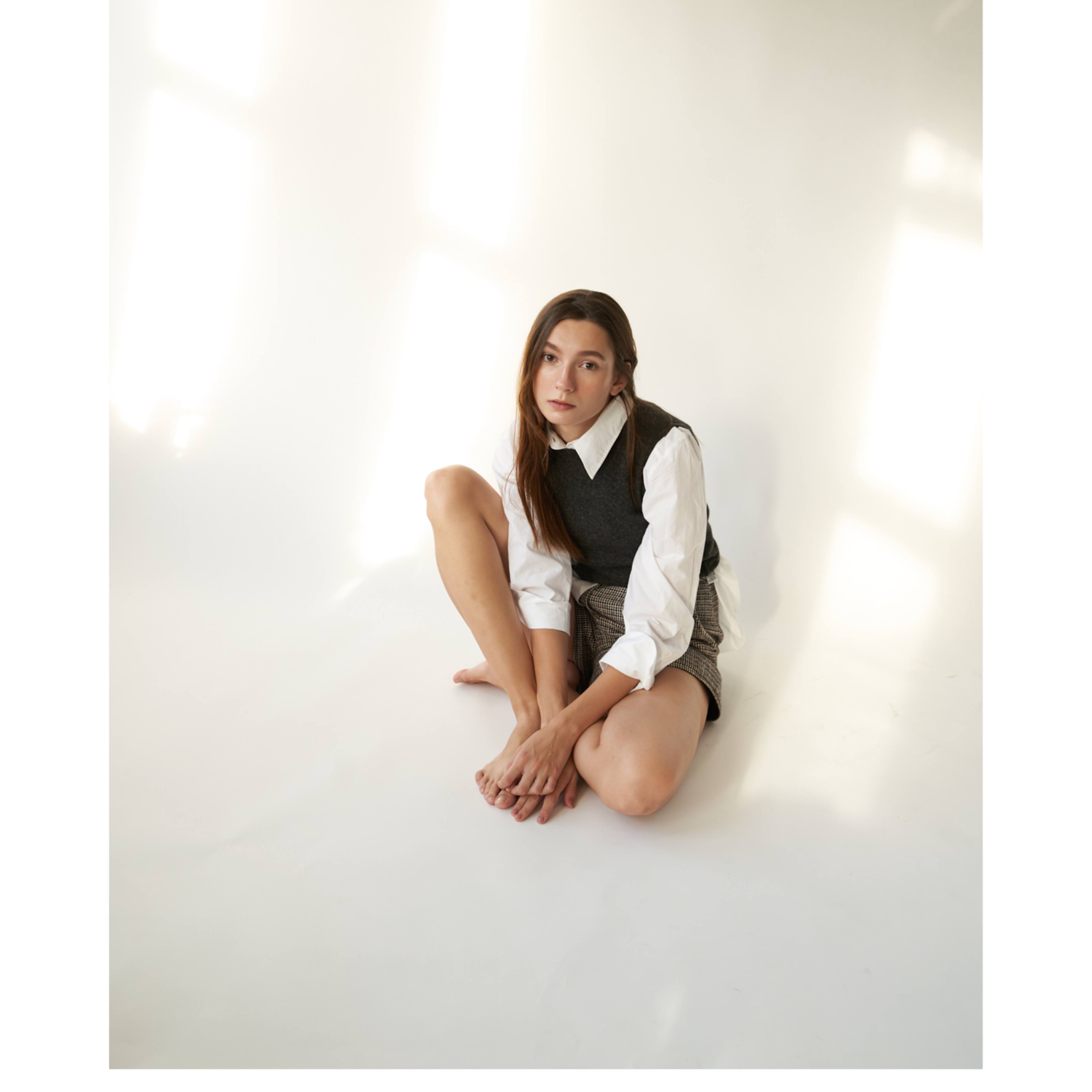 A young woman sits on the floor against a white wall, wearing a dark vest over a white shirt and patterned shorts, with sunlight streaming in from nearby windows.