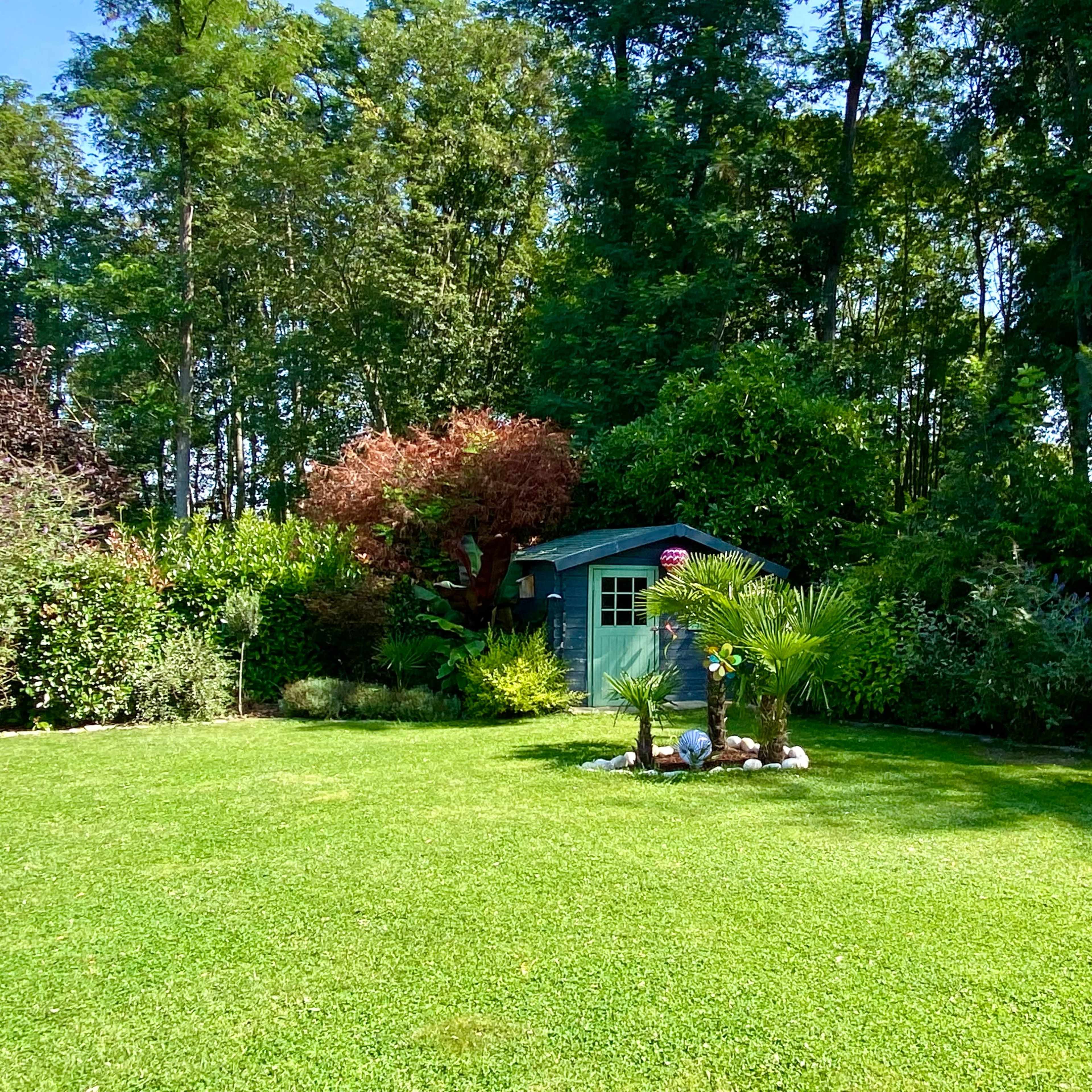 A well-maintained garden features a small green shed surrounded by various plants, bushes, and a stone statue in a serene setting.