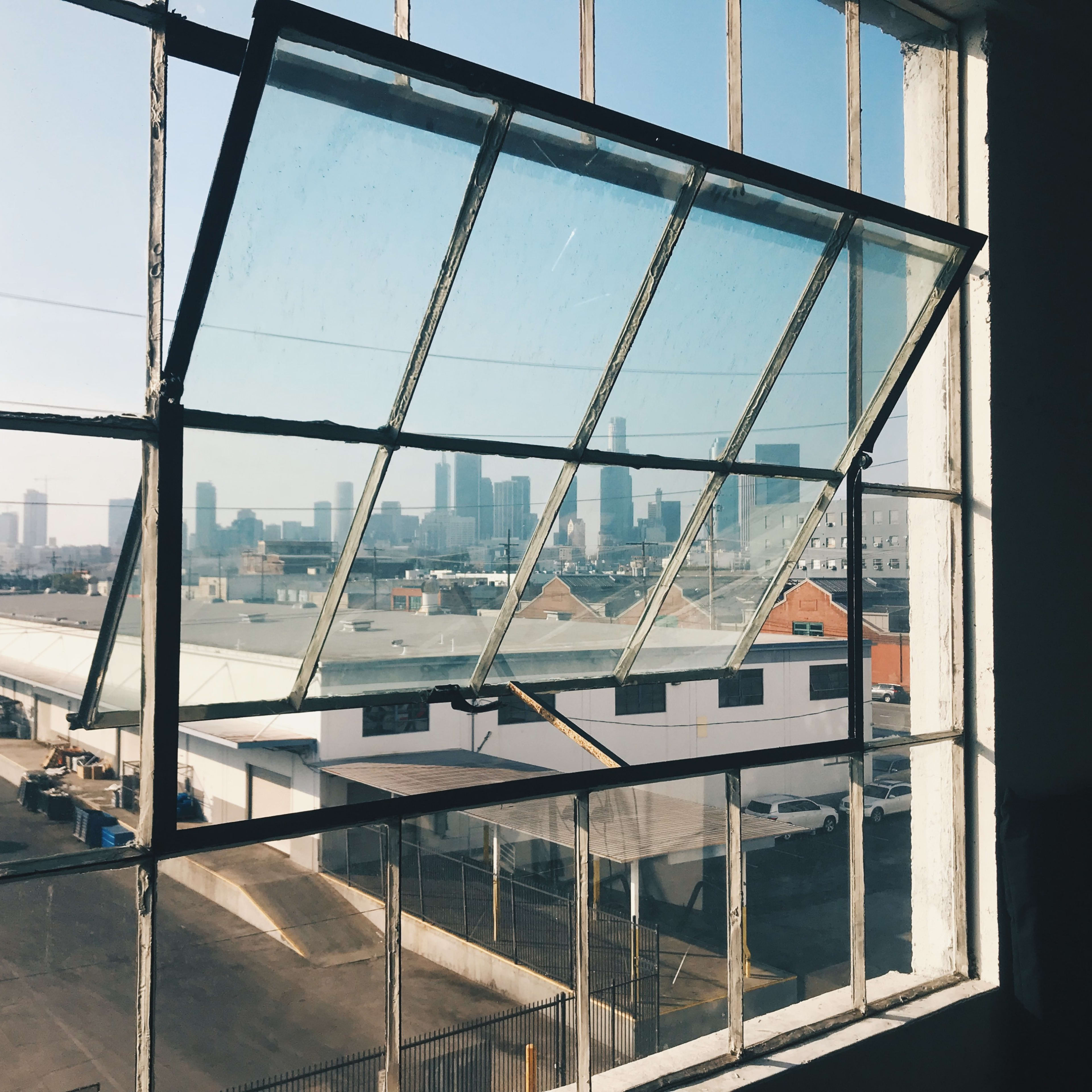 A partially open window framed by a metal structure reveals a city skyline in the distance with industrial buildings in the foreground.