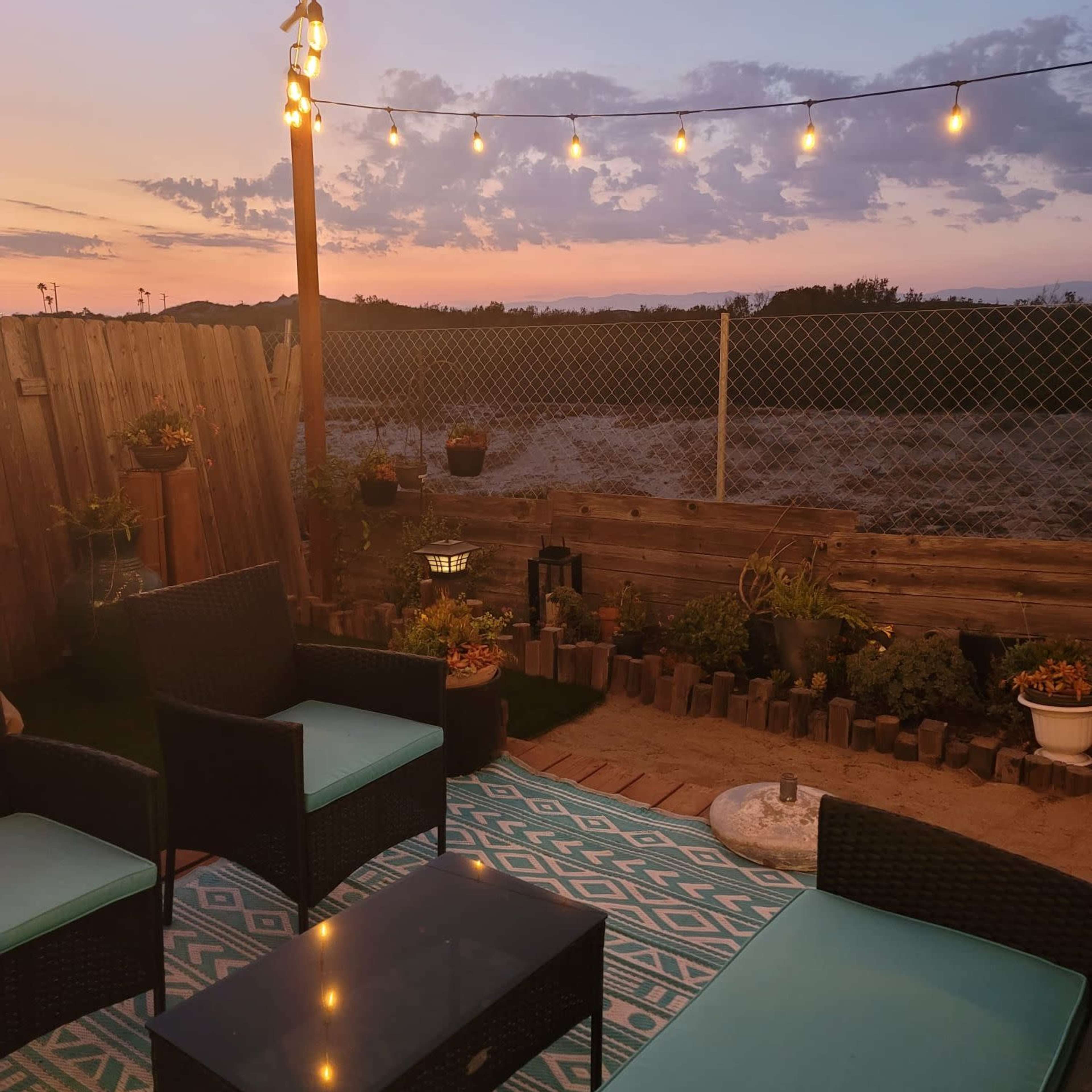 A patio area is set up with seating, a low table, and string lights overhead, overlooking a field at sunset.