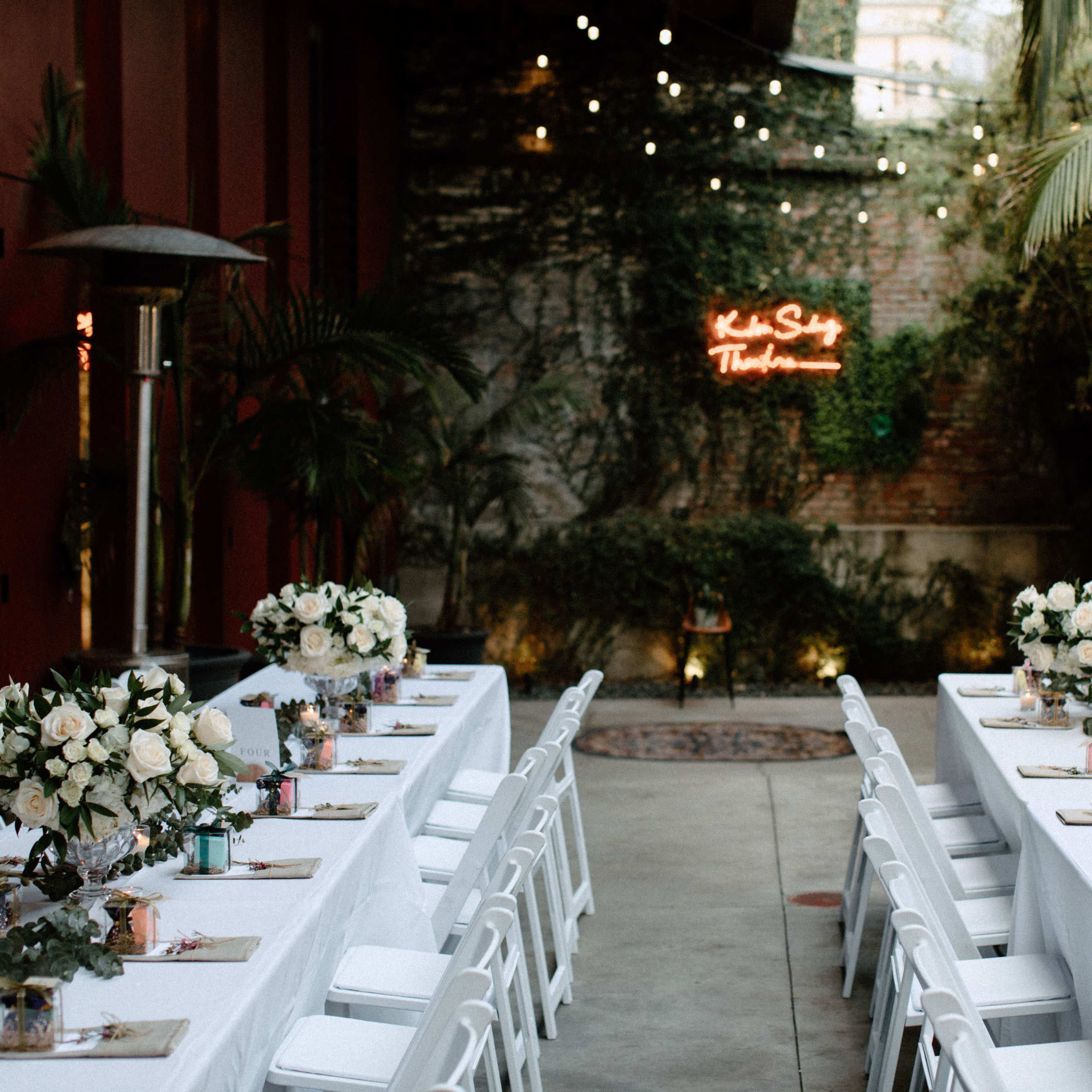 A garden venue is set up with two long tables adorned with white tablecloths and centerpieces of flowers, under string lights and a neon sign.