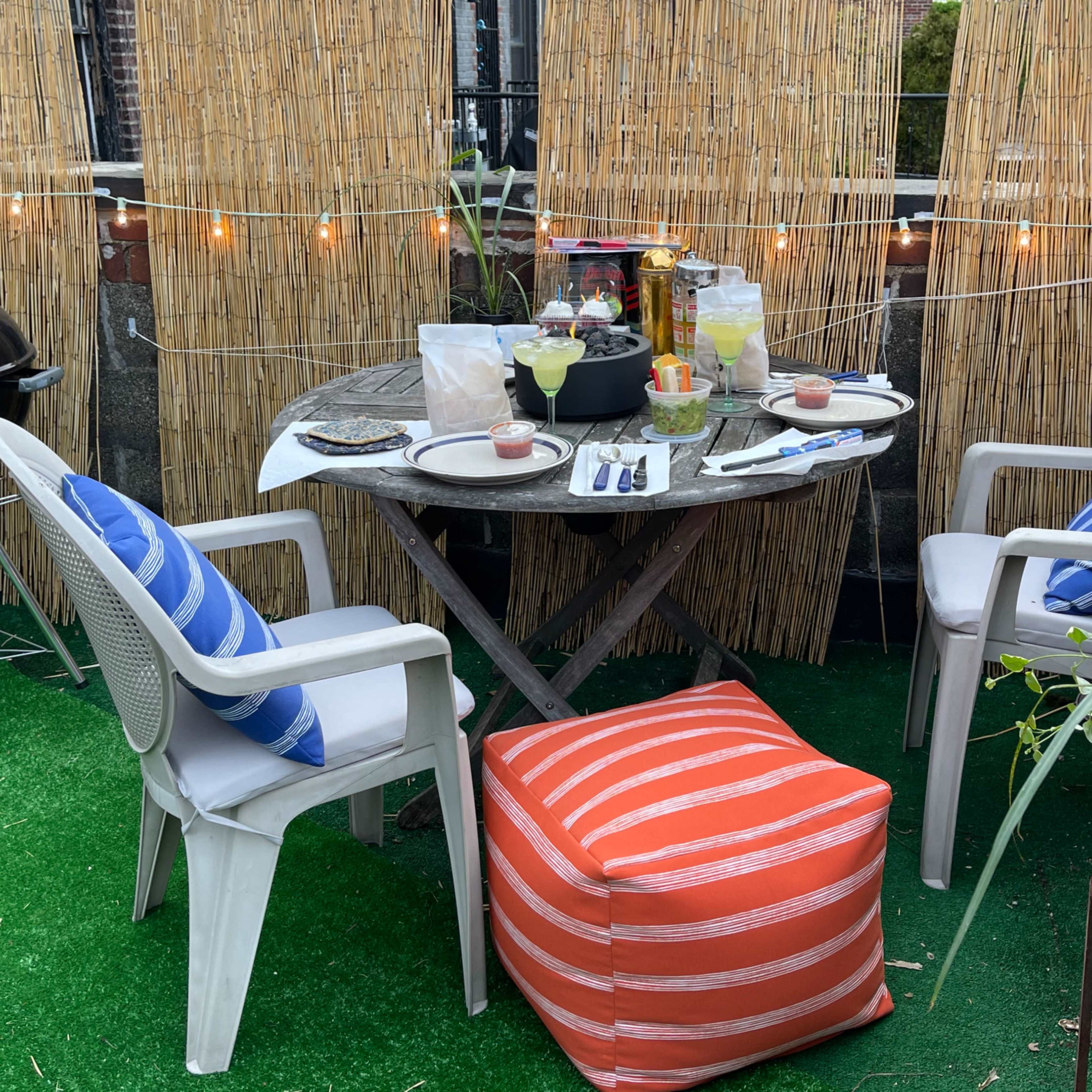 A small outdoor dining area features a round table set for a meal, surrounded by chairs and a colorful pouf, with string lights overhead and a grill nearby.