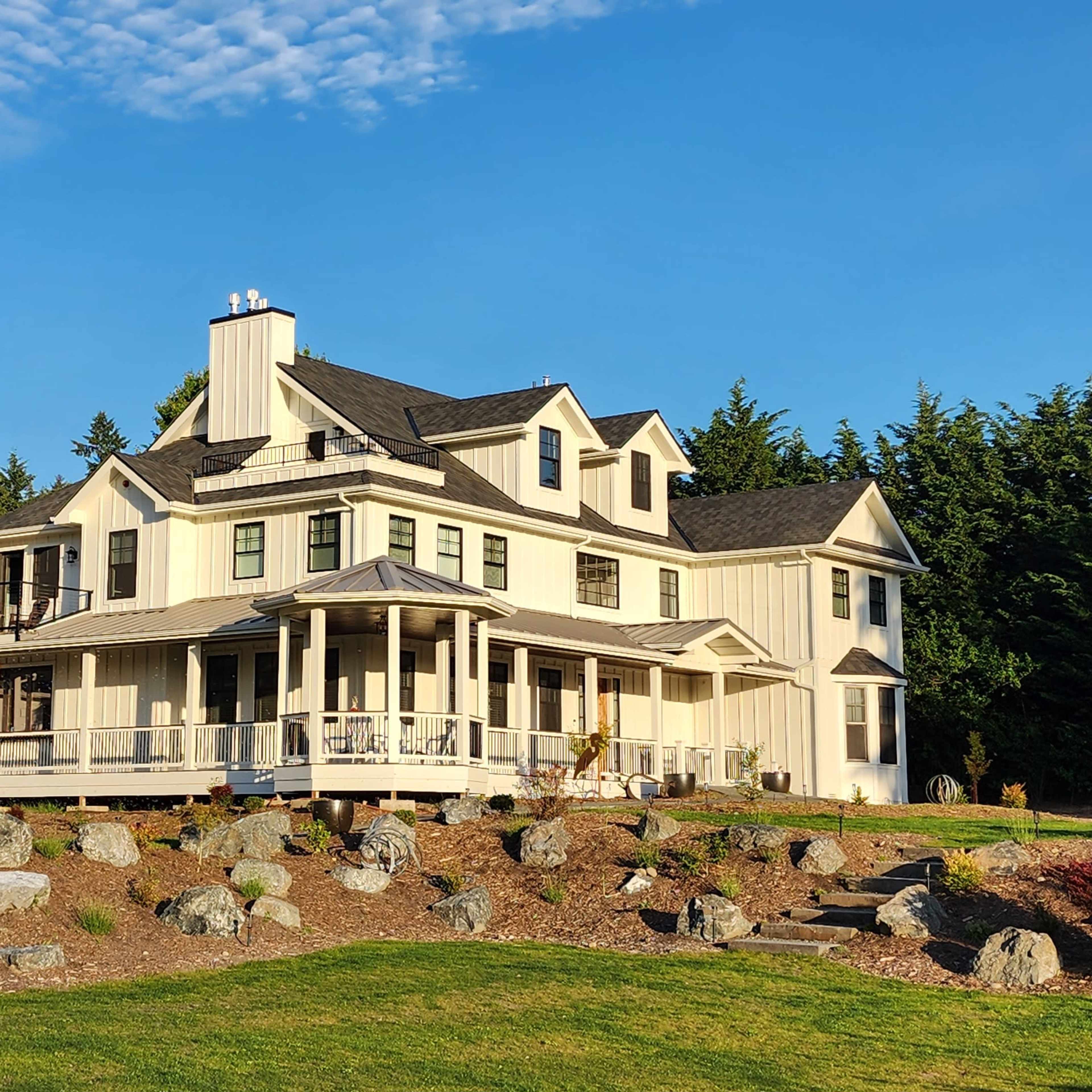 A large white house with multiple gables and a wraparound porch is surrounded by landscaped rocks and greenery on a clear day.