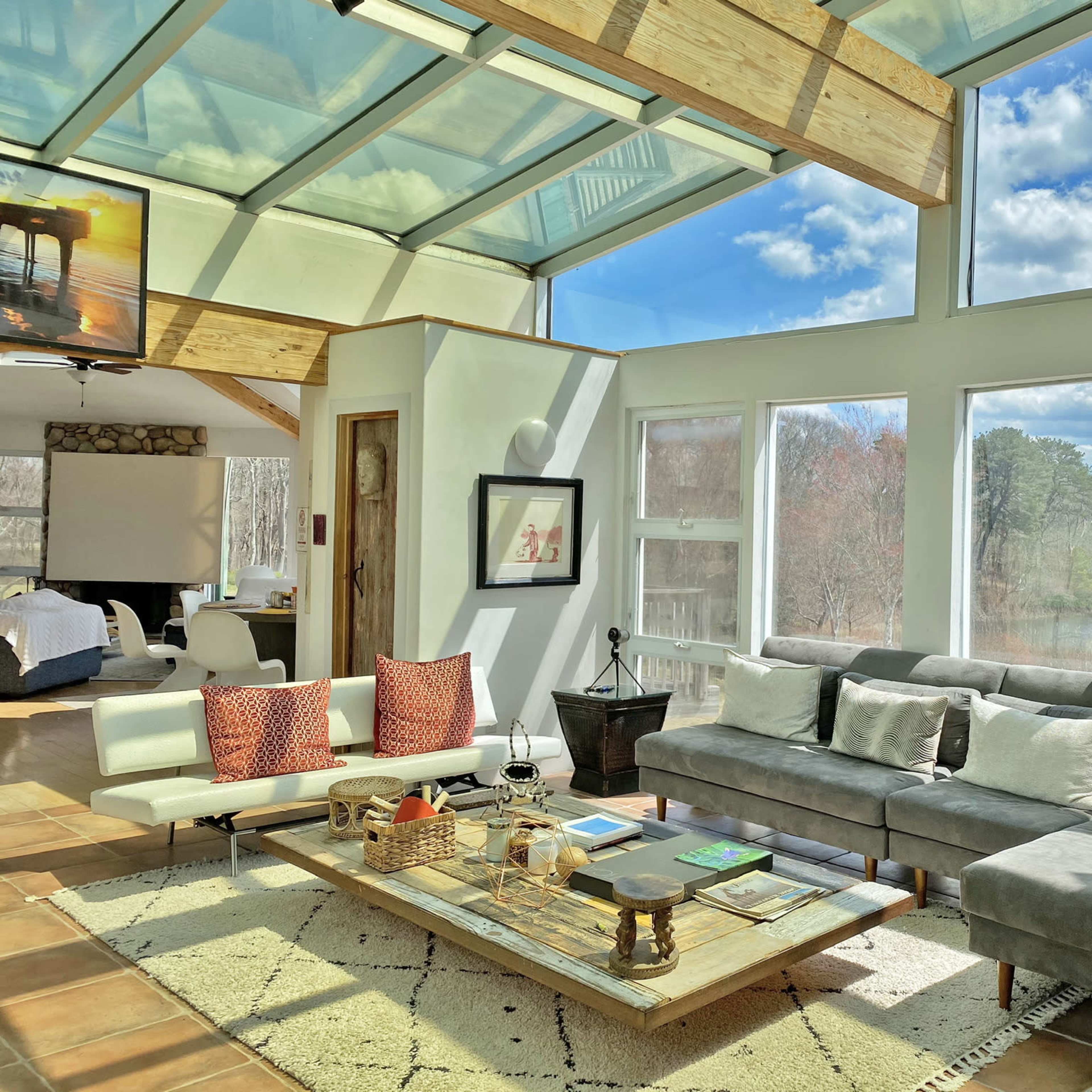 A bright and spacious living room with large windows overlooking a lake, featuring a sectional sofa, a coffee table with books, and a television mounted on the wall.
