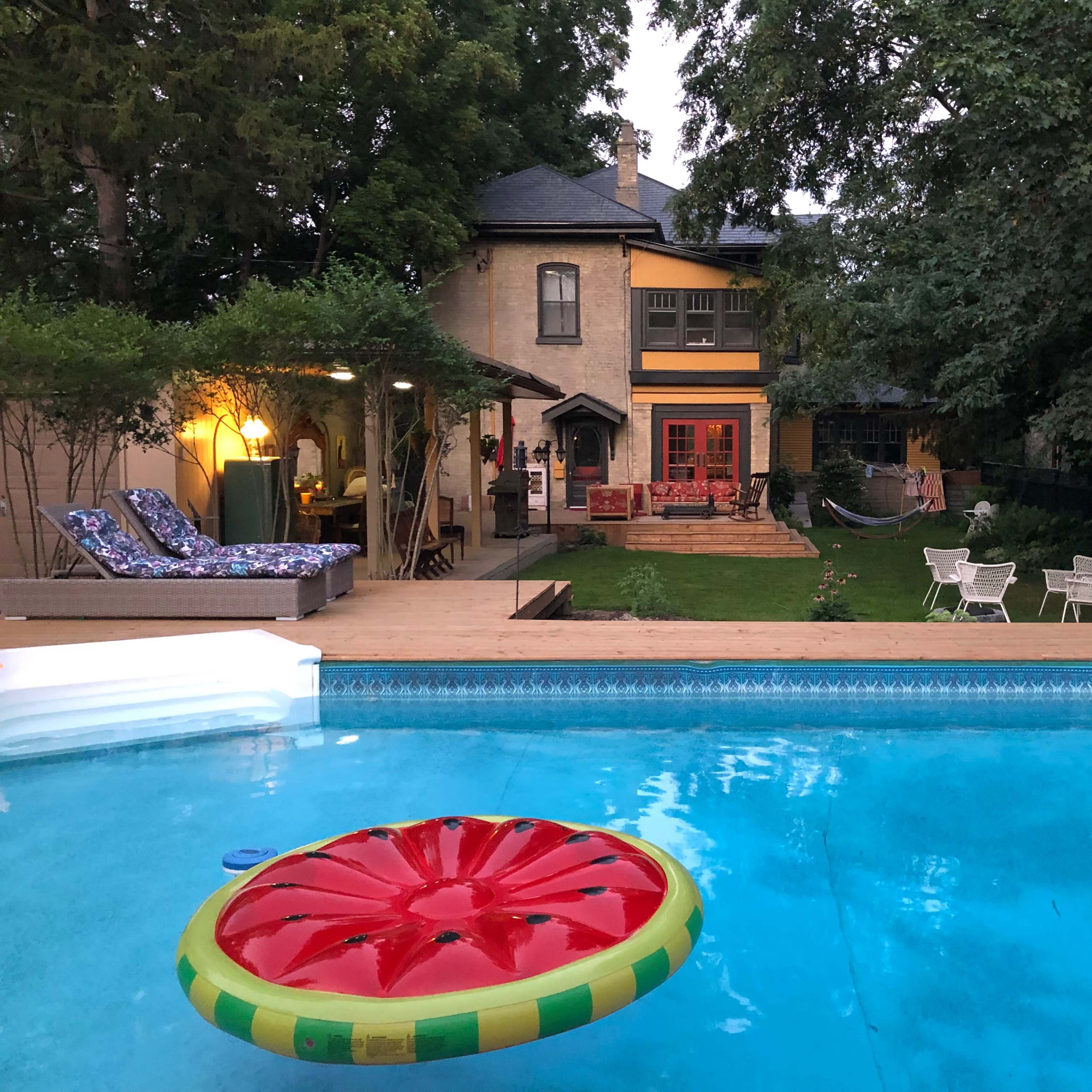 A bright red watermelon-shaped float is in the foreground of a swimming pool, with a backyard featuring a house, lounge chairs, and a hammock beyond.