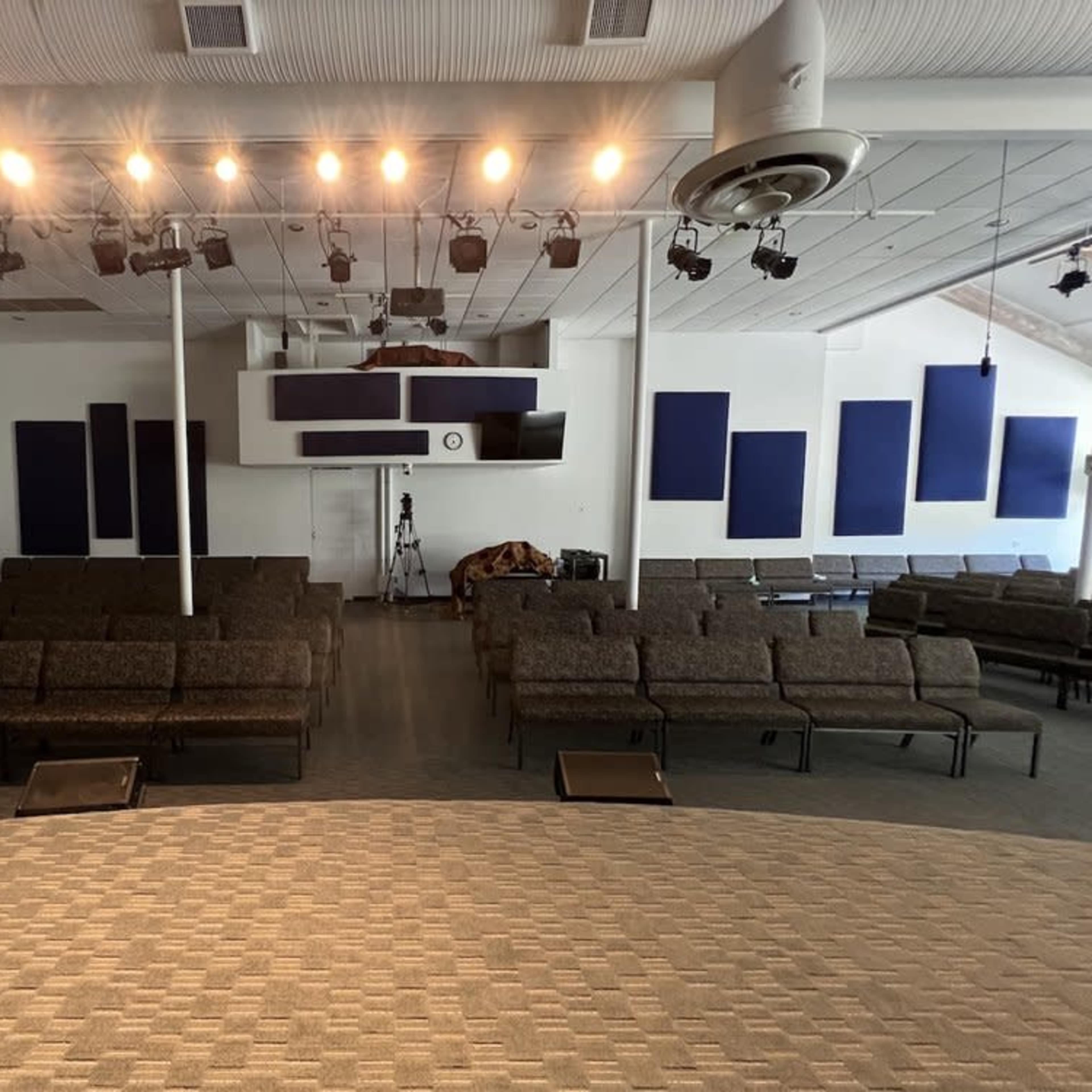 An empty auditorium with rows of chairs facing a stage, featuring overhead lights and sound equipment.