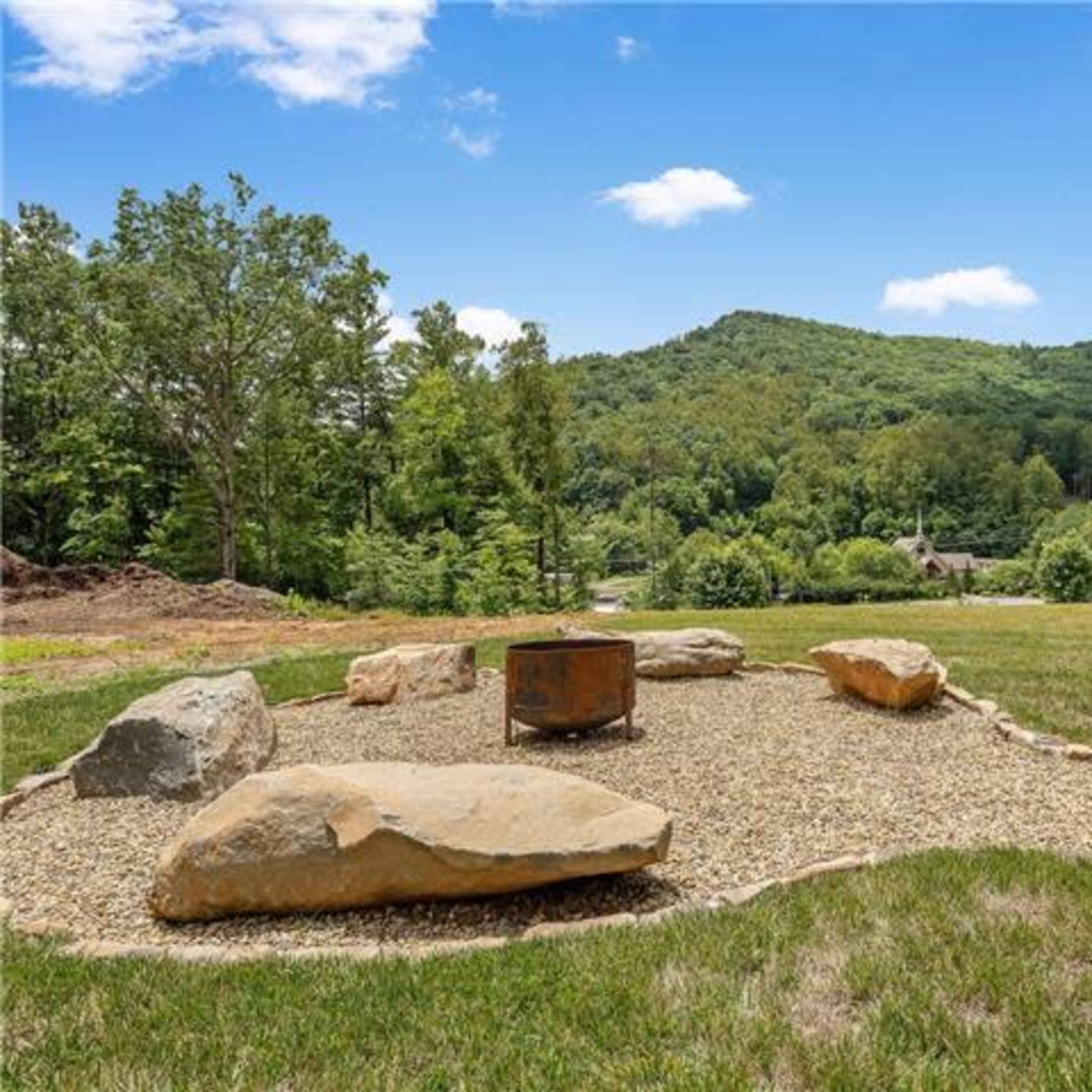A fire pit surrounded by large rocks is set in a gravel landscape with green hills in the background.