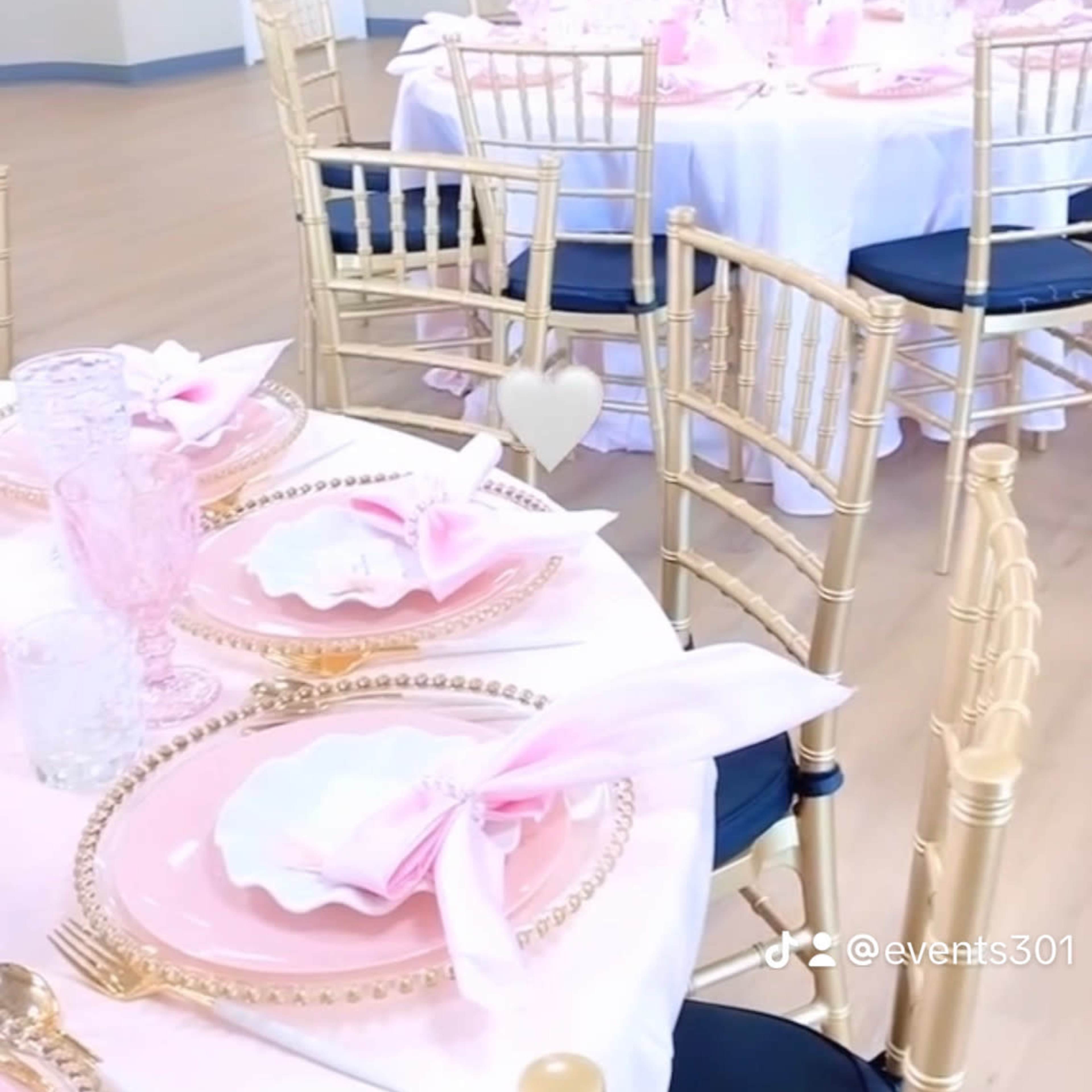 A decorated event space with round tables set with pink tableware and gold chairs.