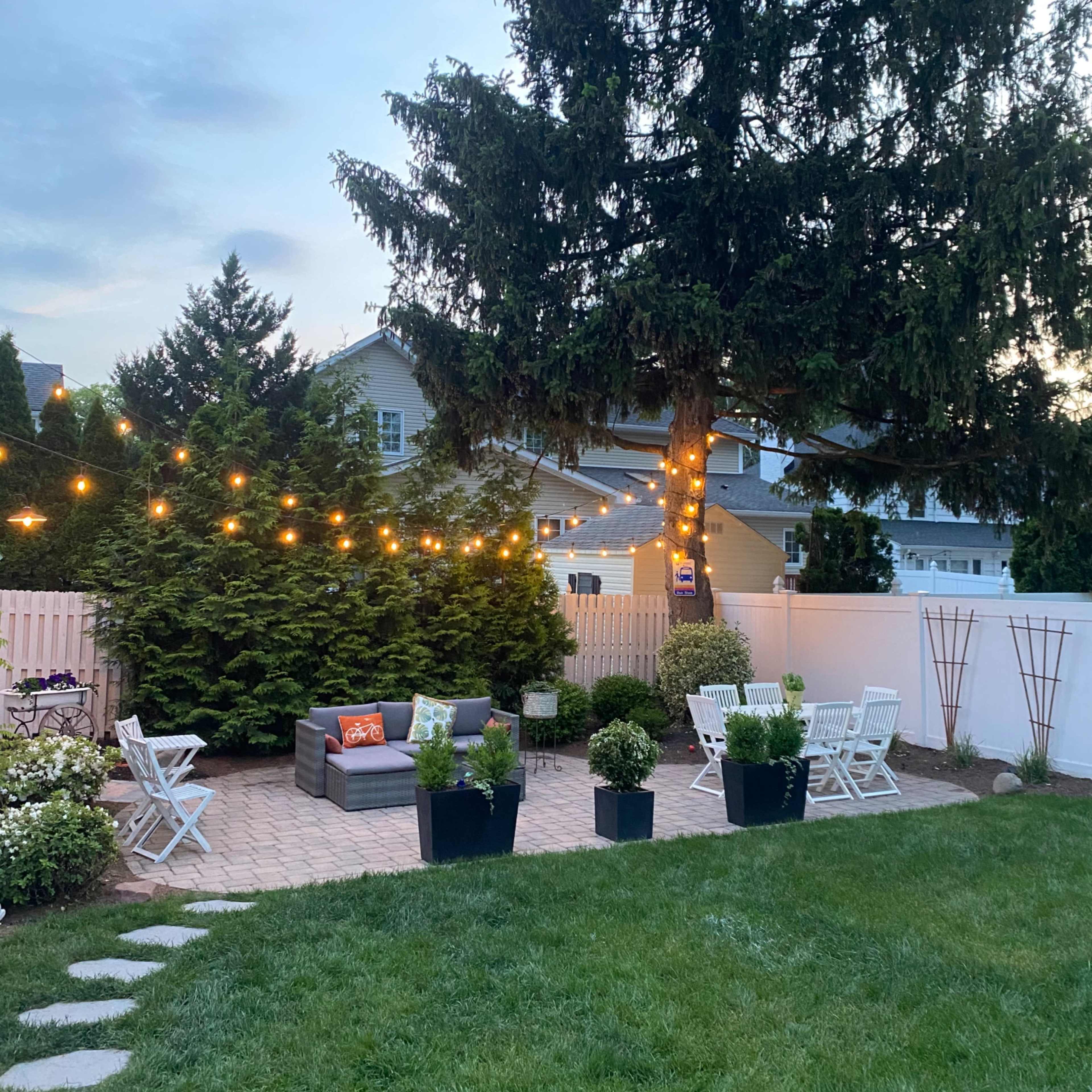 A backyard features a seating area with string lights above and potted plants arranged around a grassy lawn.