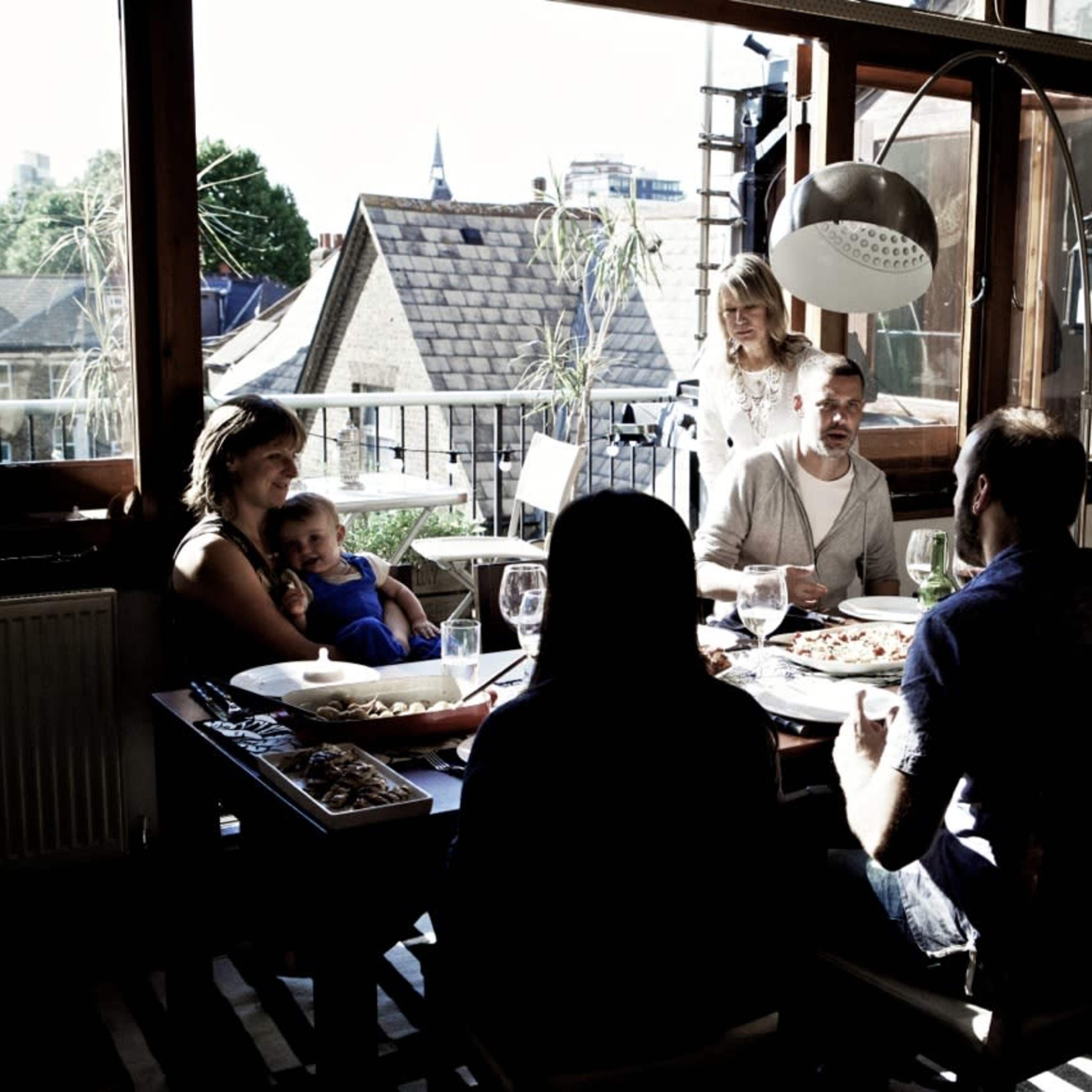 A group of six people sits around a table in a sunlit room, sharing food and conversation.