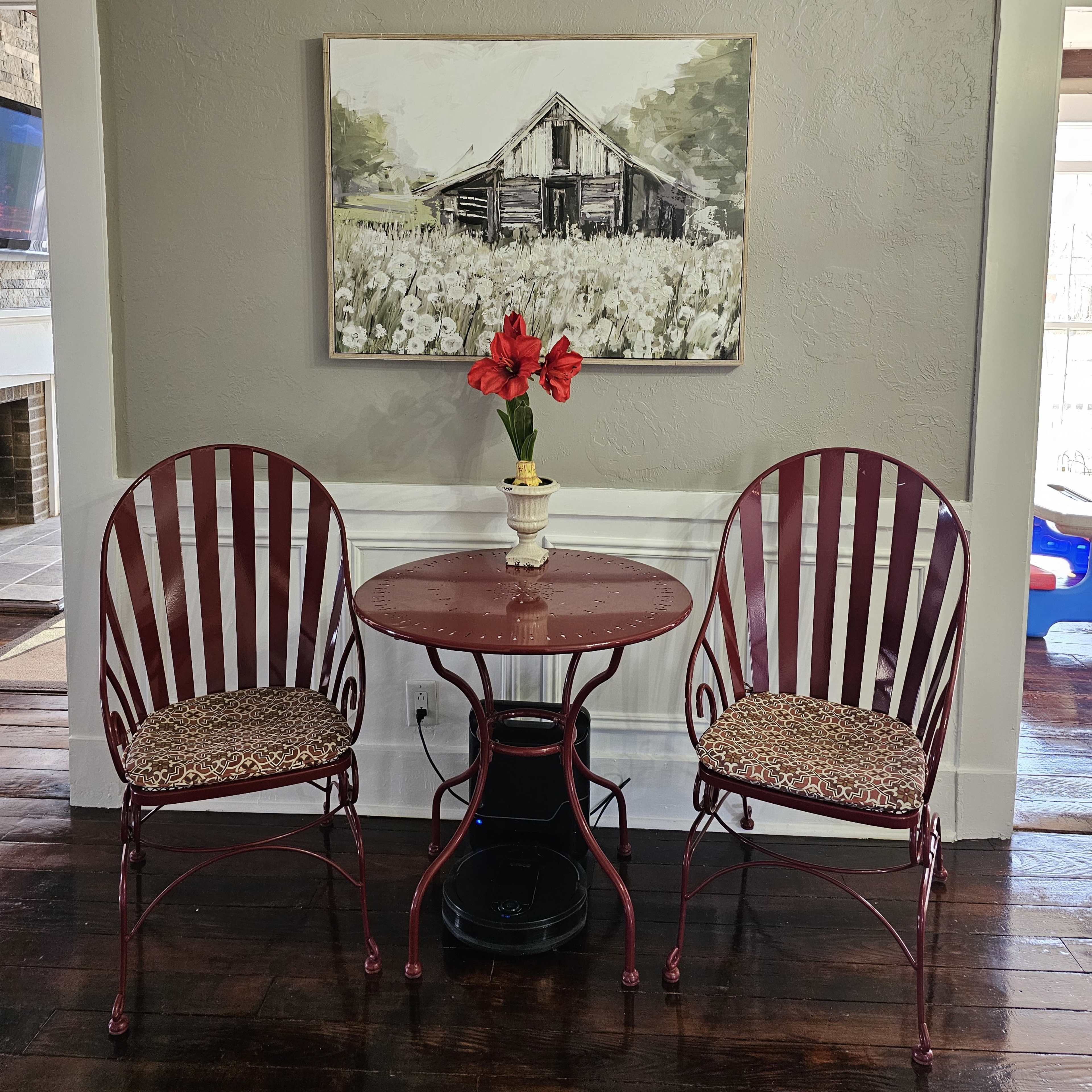 A small round table with two chairs is positioned against a gray wall, featuring a floral centerpiece and a painting of a barn above it.