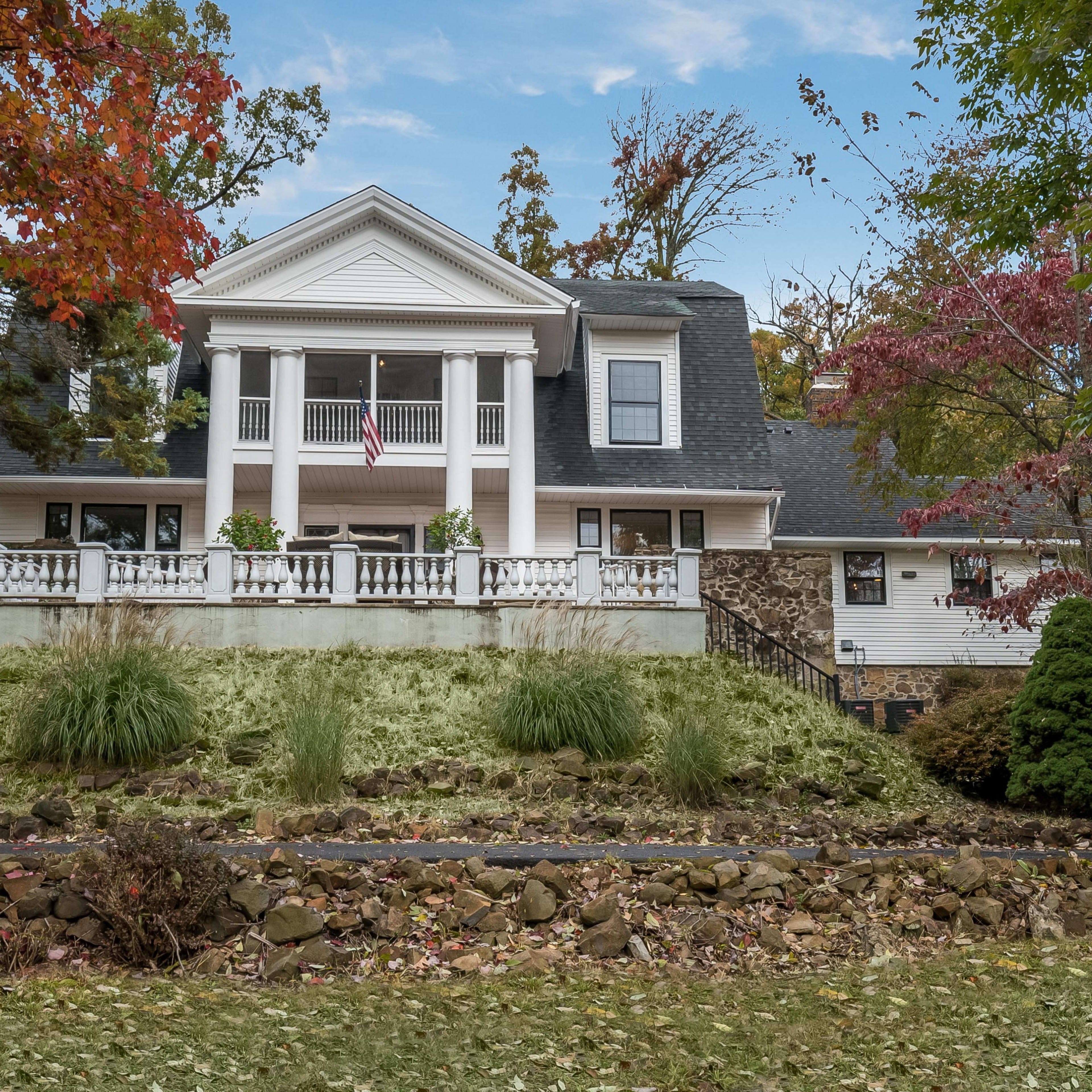 A two-story house with a large front porch, situated on a grassy hill surrounded by trees and autumn foliage.