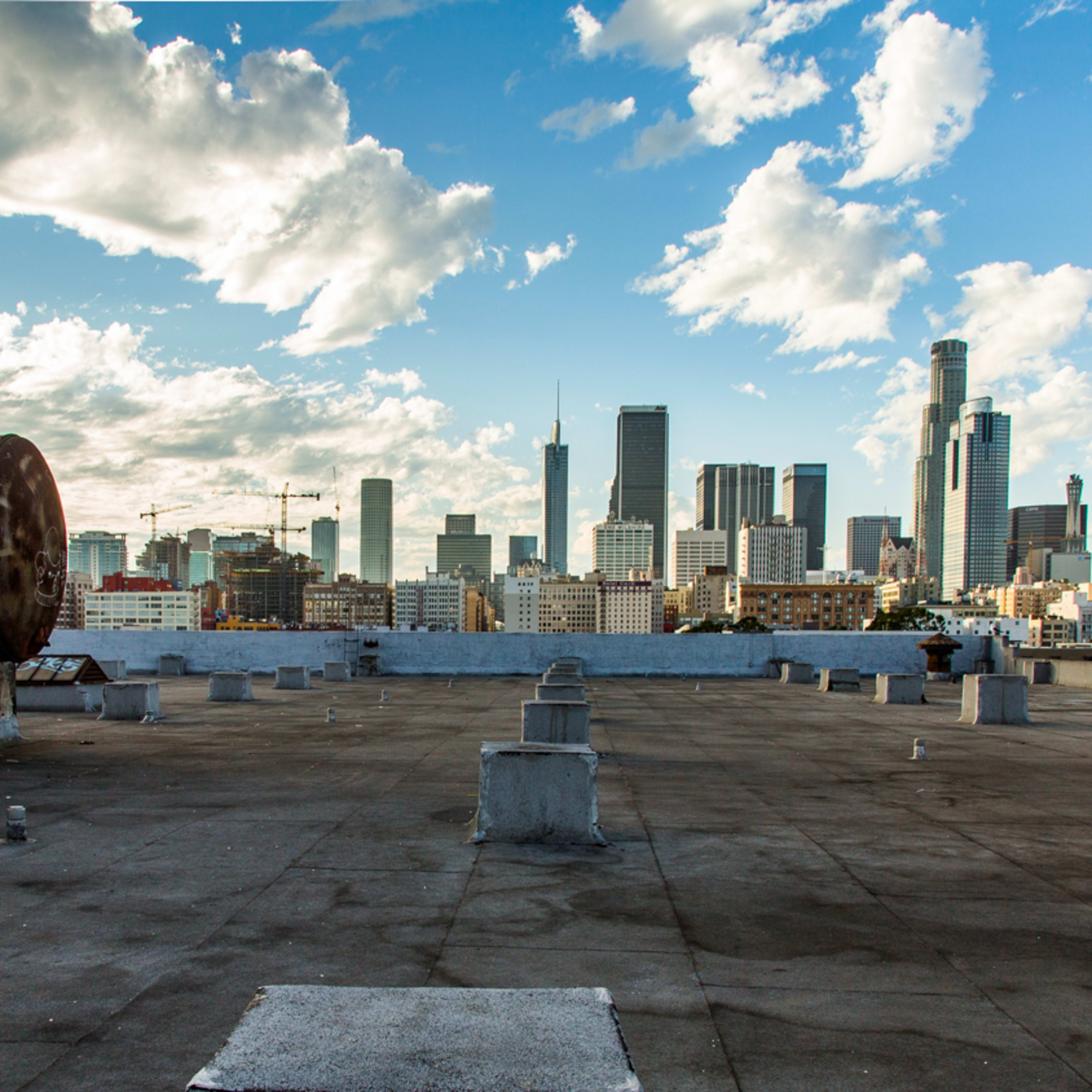 A rooftop with three large, rusty tanks overlooking a city skyline filled with tall buildings under a partly cloudy sky.