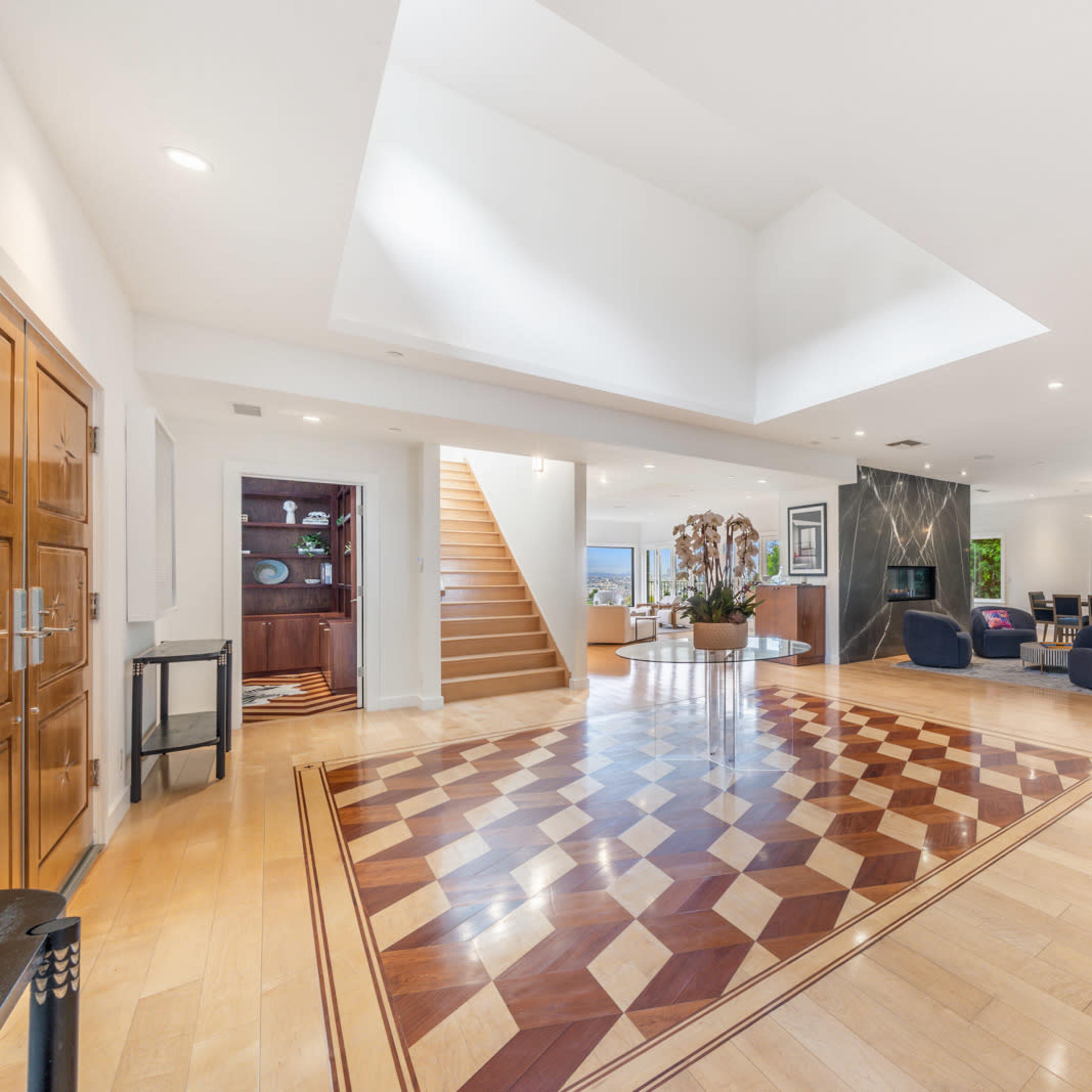 A spacious, modern living area with a patterned wooden floor, a staircase in the background, and large windows providing natural light.