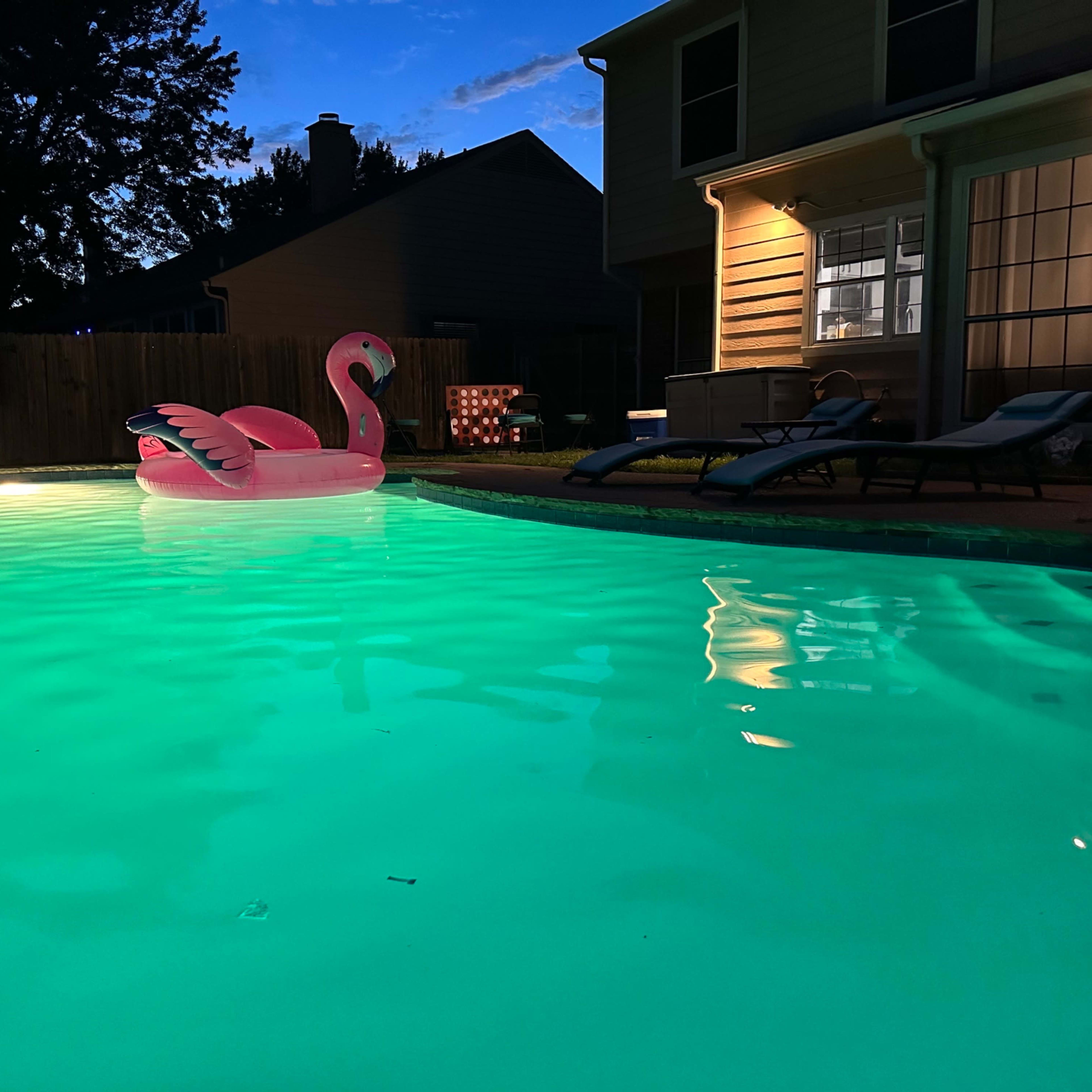 A pink flamingo float rests in a lit turquoise swimming pool beside a house at twilight.
