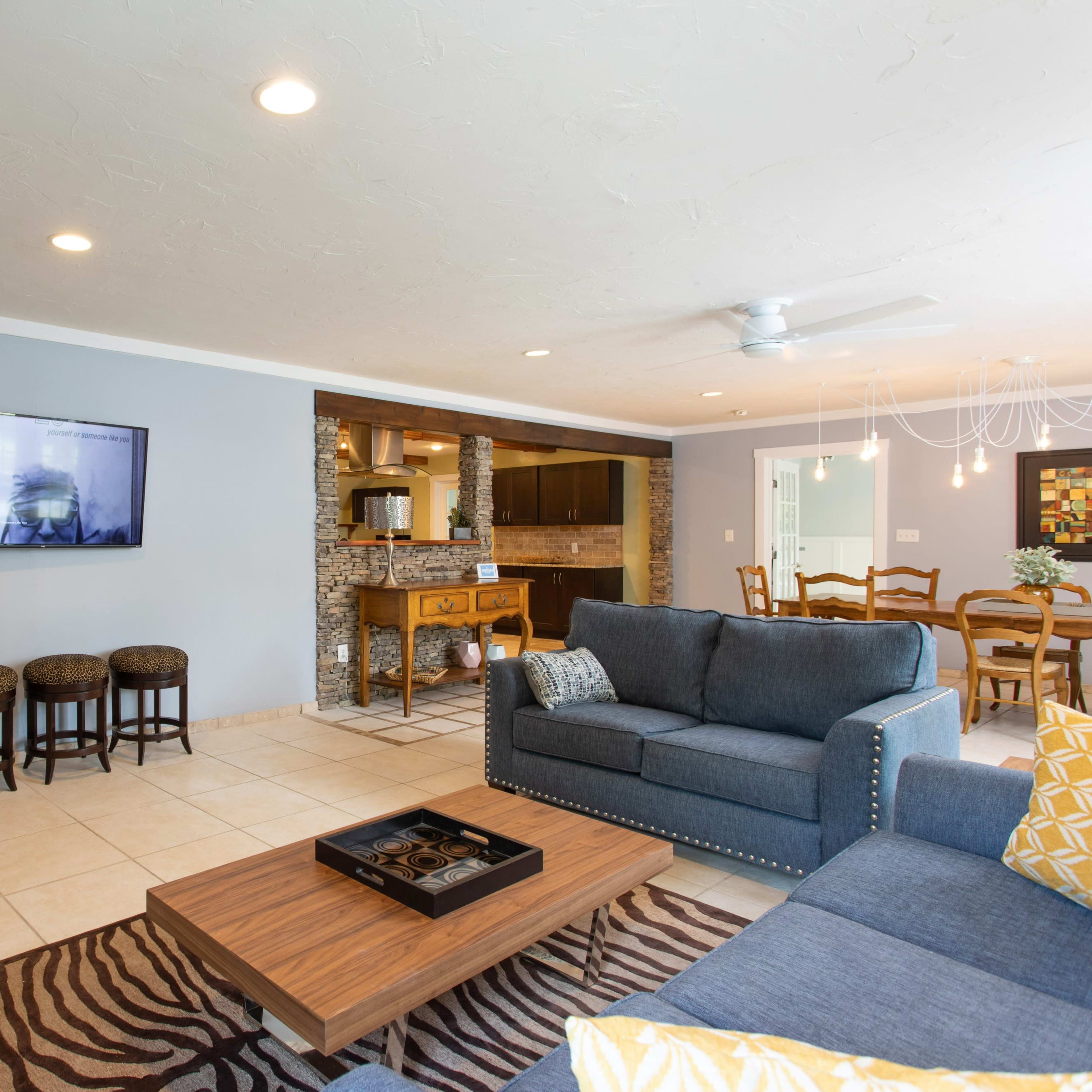 A modern living room with a blue sofa, a wooden coffee table, and a wall-mounted television, adjacent to a dining area and kitchen.